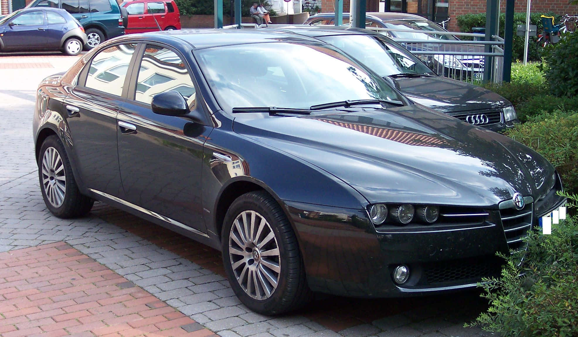Alfa Romeo 159 桌布 壁紙 — 一輛時尚的深藍色 Alfa Romeo 房車停泊在磚砌停車場，背景有綠化及其他車輛。