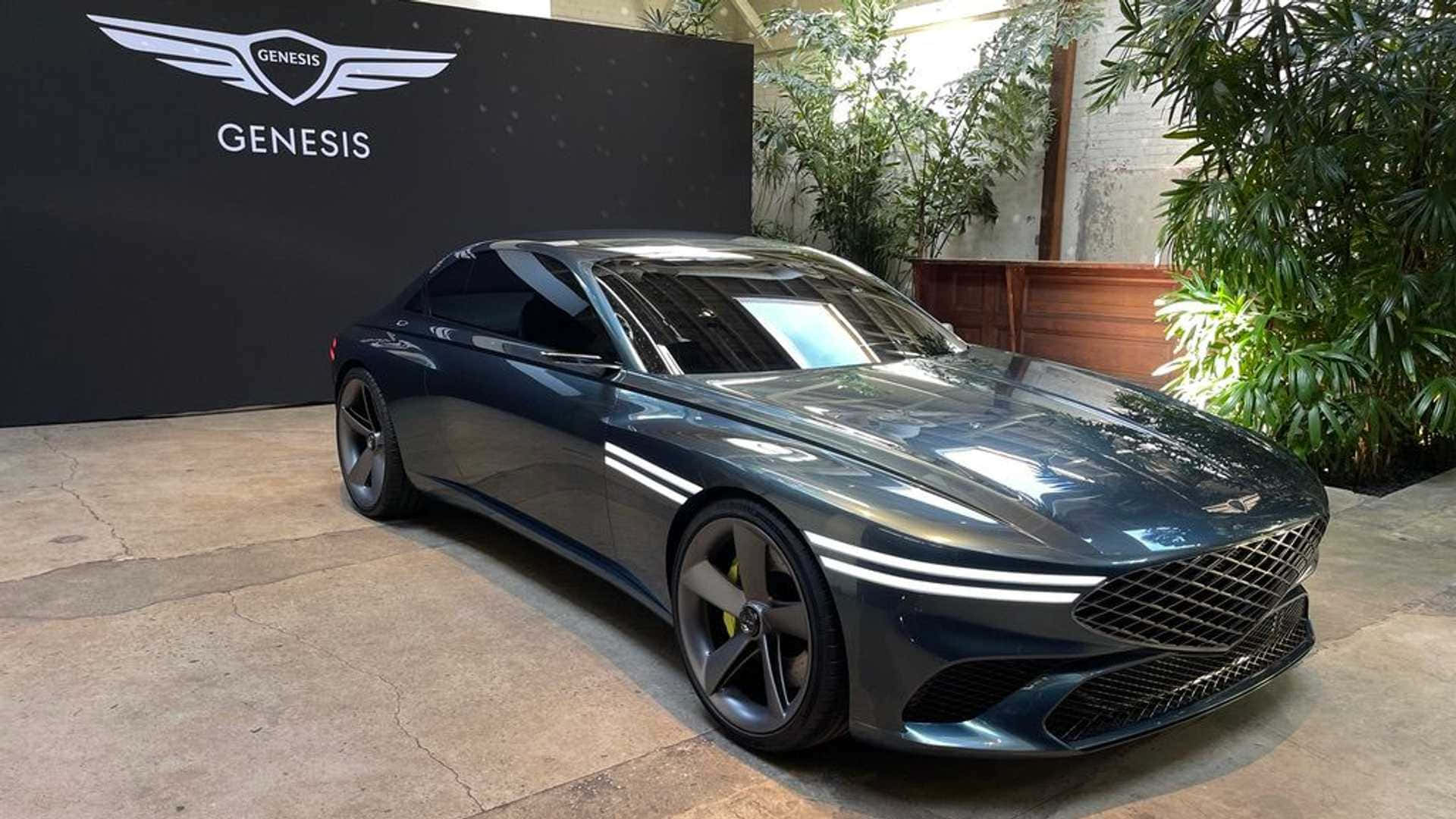 Genesis X Concept 桌布 壁紙 — 一輛時尚的深藍色 Genesis 跑車在明亮的燈光下，在品牌牆和茂密綠植旁閃閃發光。