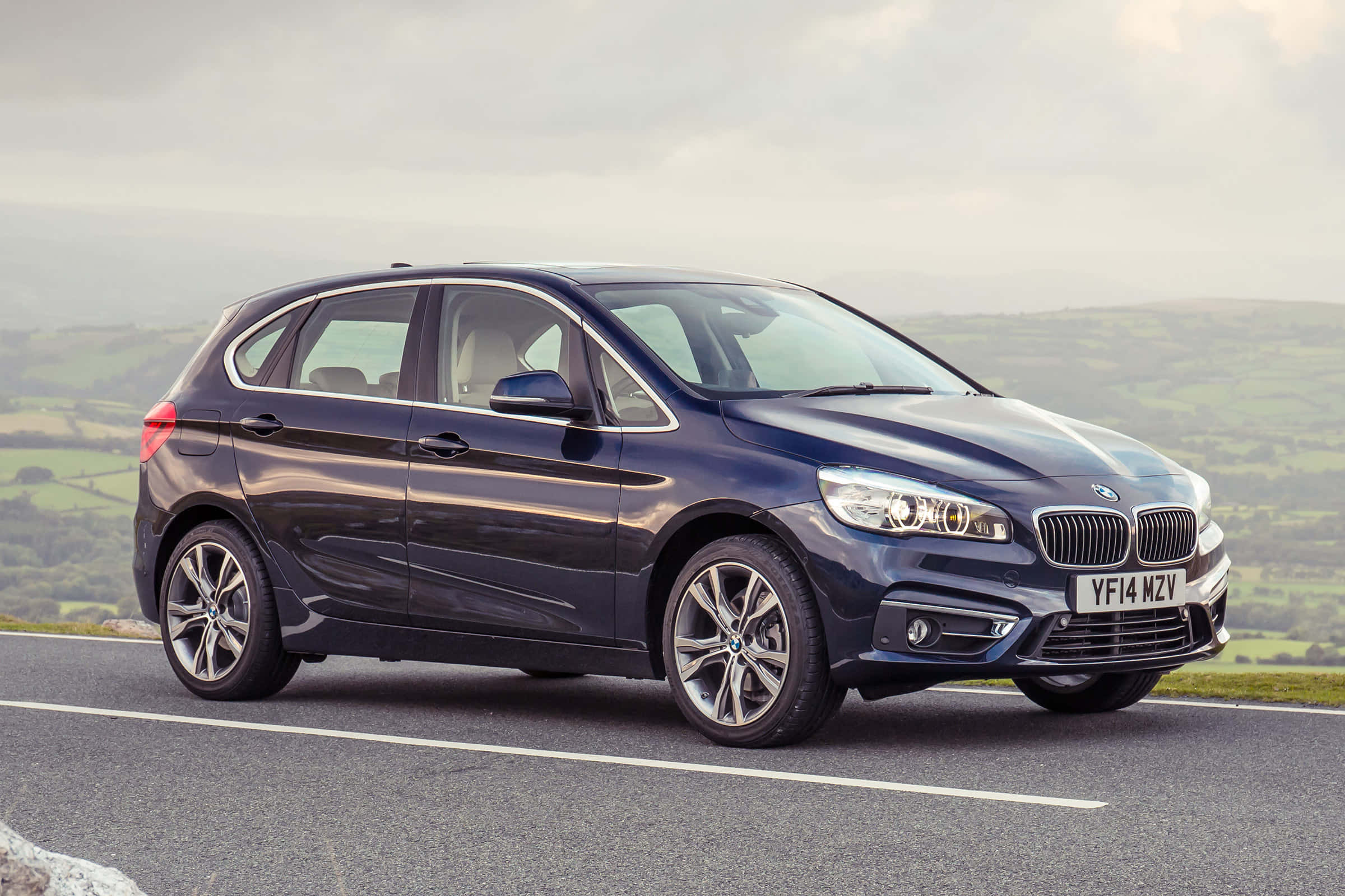 Bmw 2 Series 桌布 壁紙 — 一輛時尚的深藍色BMW 2系Active Tourer在陰天下行駛在蜿蜒的鄉村道路上，兩旁是起伏的綠色山丘。