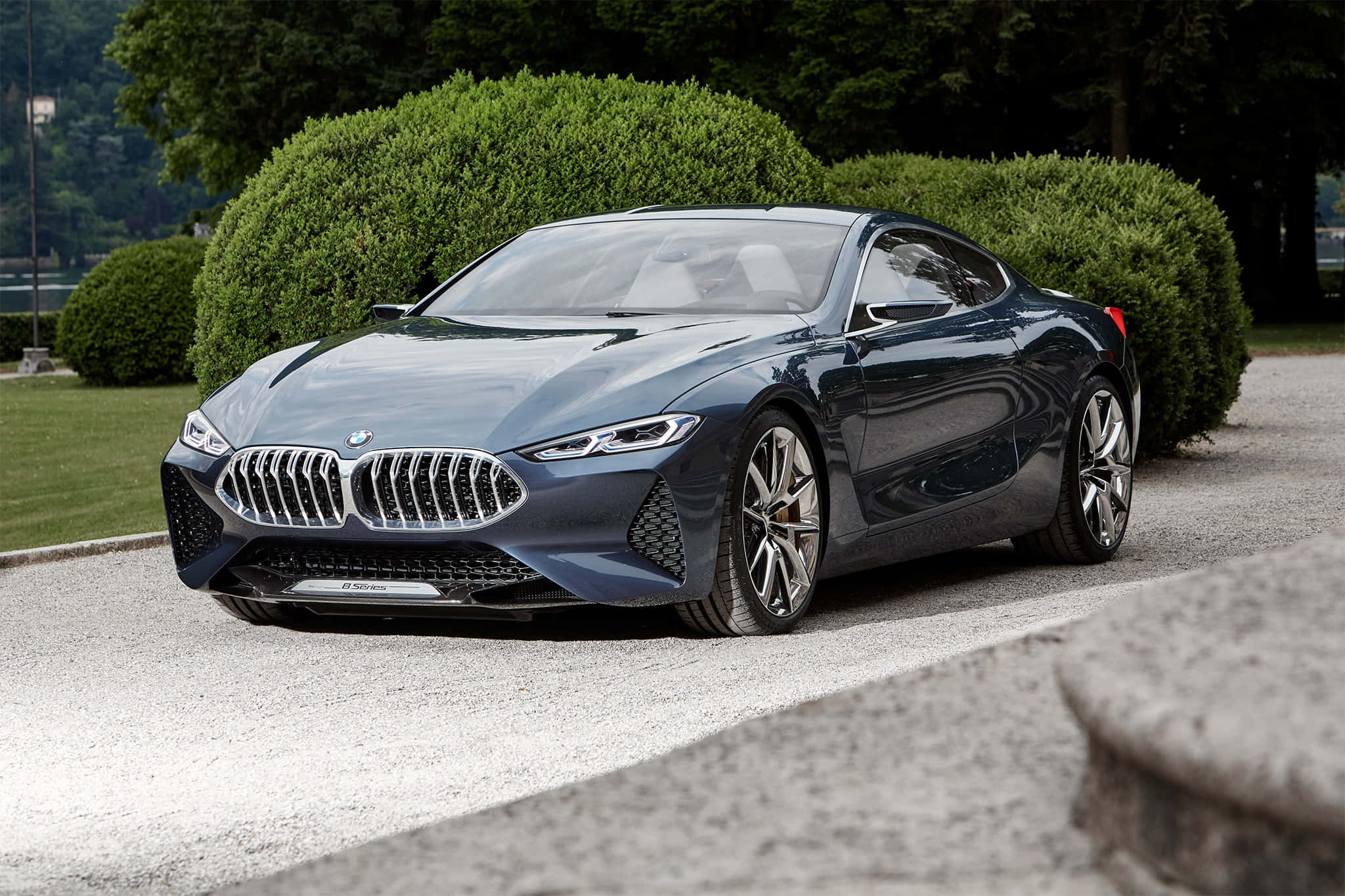 Bmw 8 Series 桌布 壁紙 — 一輛時尚的深藍色BMW概念車在日光下閃耀，停泊在碎石路上，旁邊是修剪整齊的綠色樹籬。