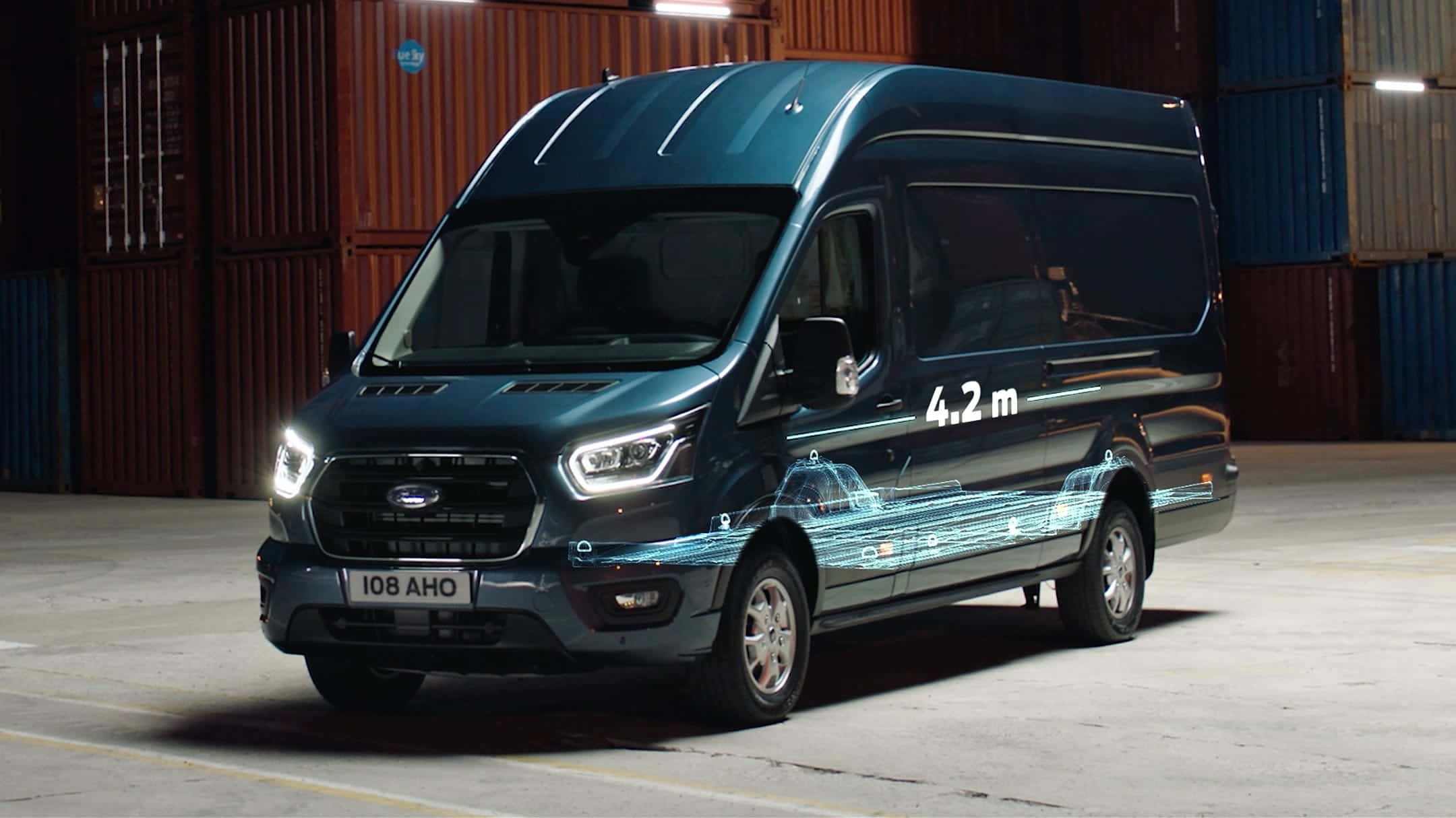Ford Transit 桌布 壁紙 — 一輛時尚的深藍色福特 Transit 客貨車，車側帶有發光的數碼線條和「4.2 m」字樣，停放在一個有貨櫃的倉庫中。