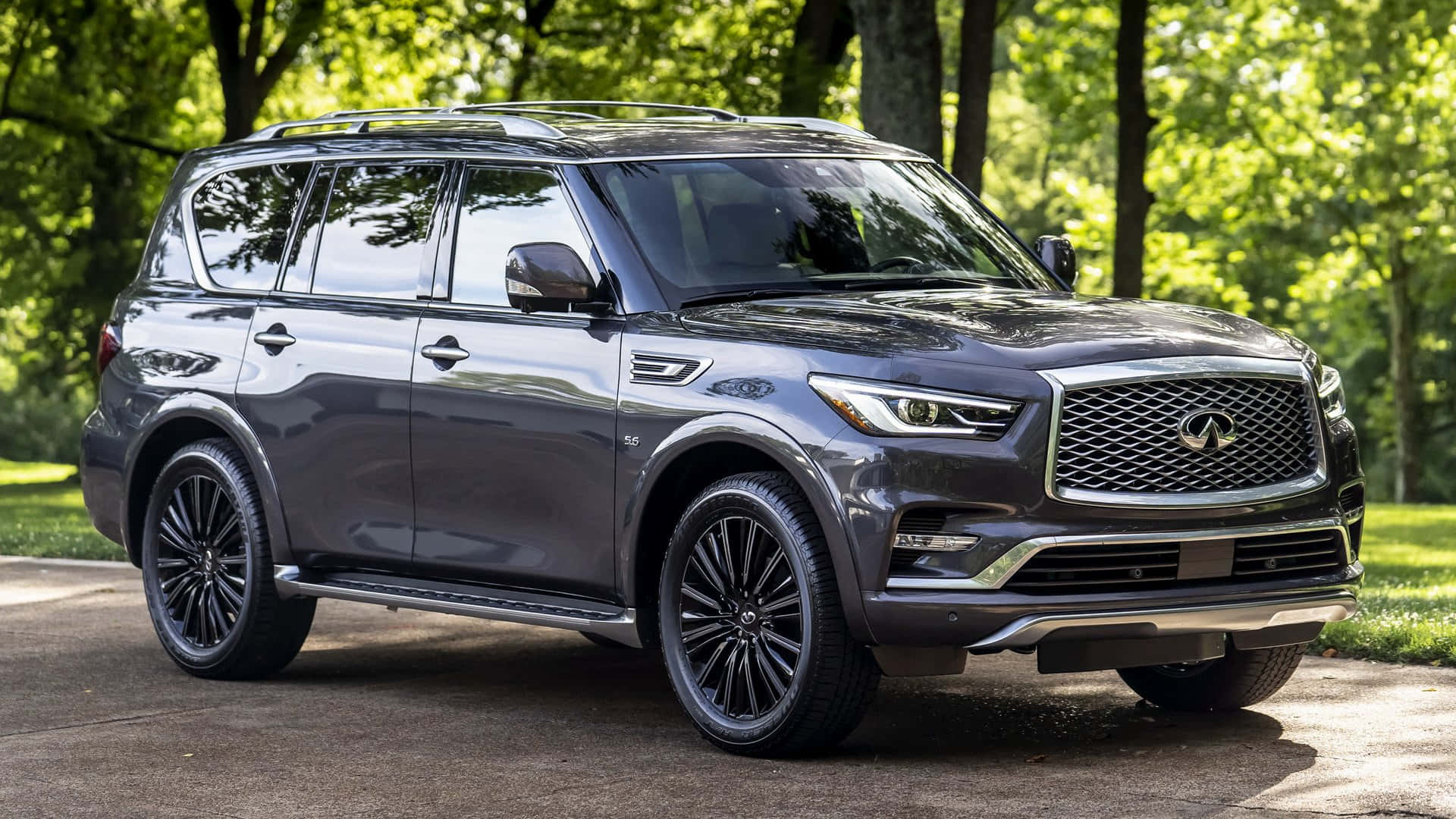 Infiniti Qx80 桌布 壁紙 — 一輛時尚的灰色 Infiniti QX80 SUV 停在鋪砌的小路上，周圍是茂密的綠樹，在明亮的日光下。