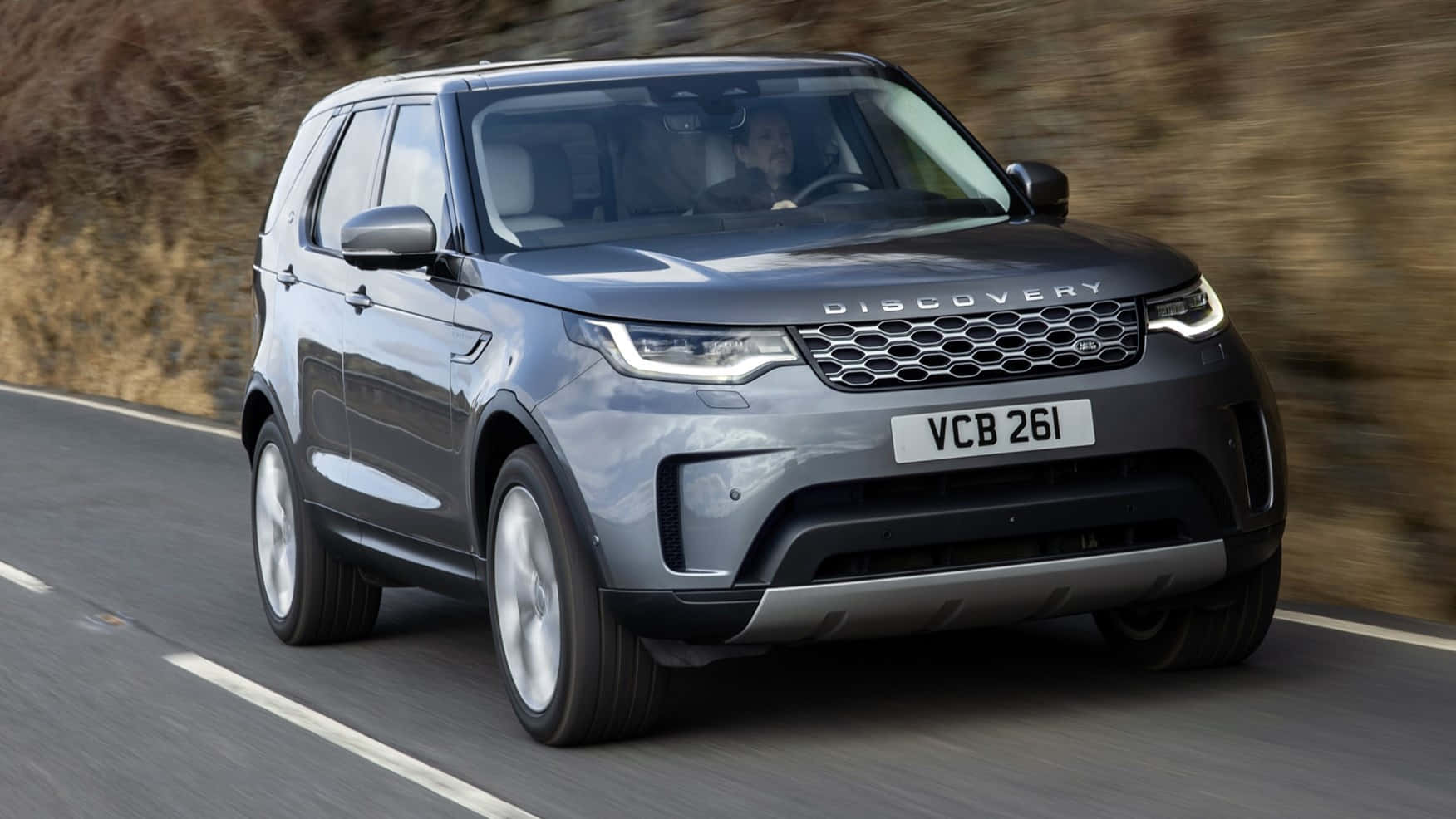 Land Rover Discovery 桌布 壁紙 — 一輛時尚的灰色 Land Rover Discovery 在蜿蜒的道路上快速行駛，模糊的土色山坡在移動中。