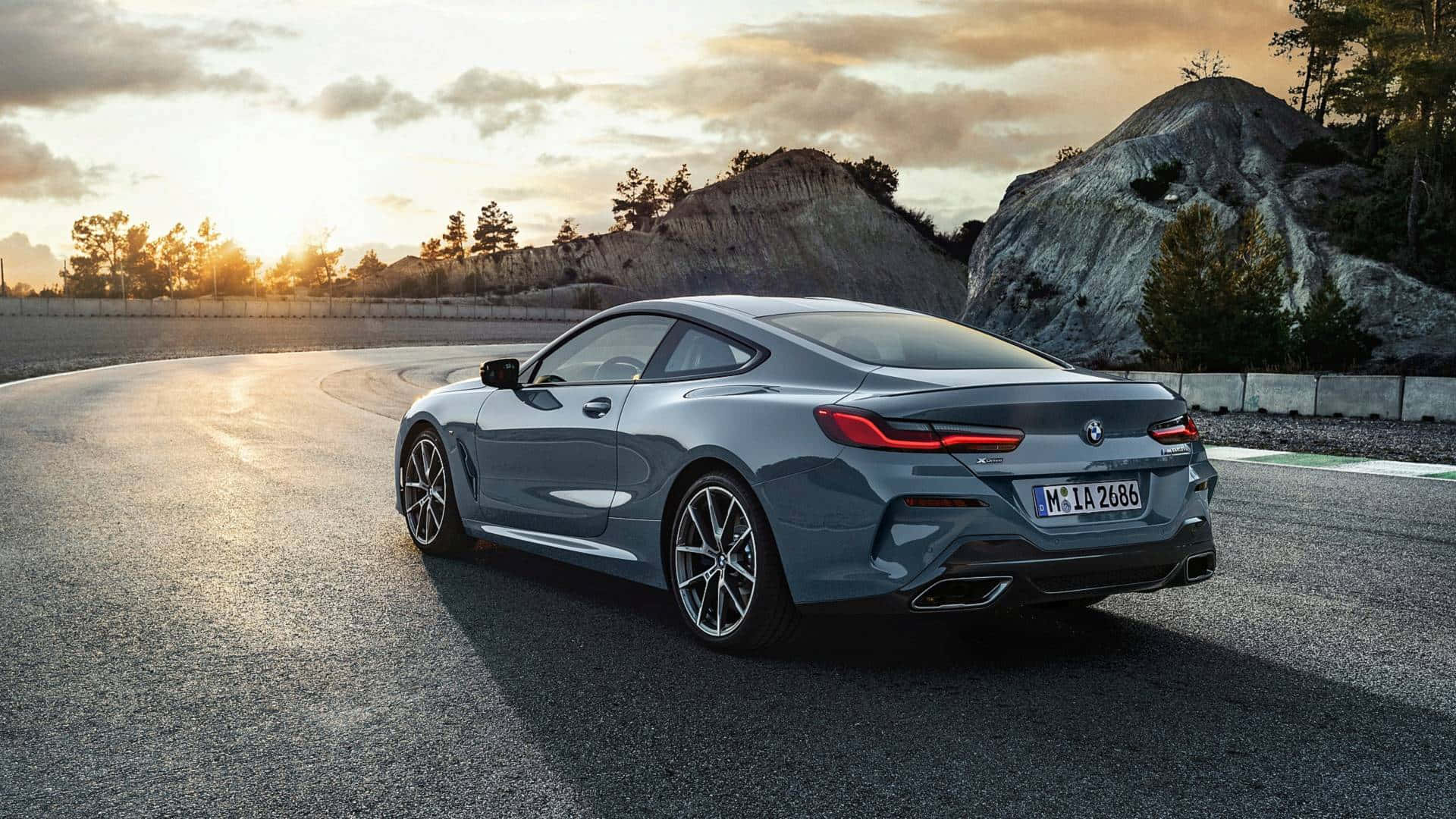 Bmw 8 Series 桌布 壁紙 — 一輛時尚的灰色BMW 8系轎跑車在日落時分沿著蜿蜒的瀝青道路行駛，背景是岩石丘陵和樹木。