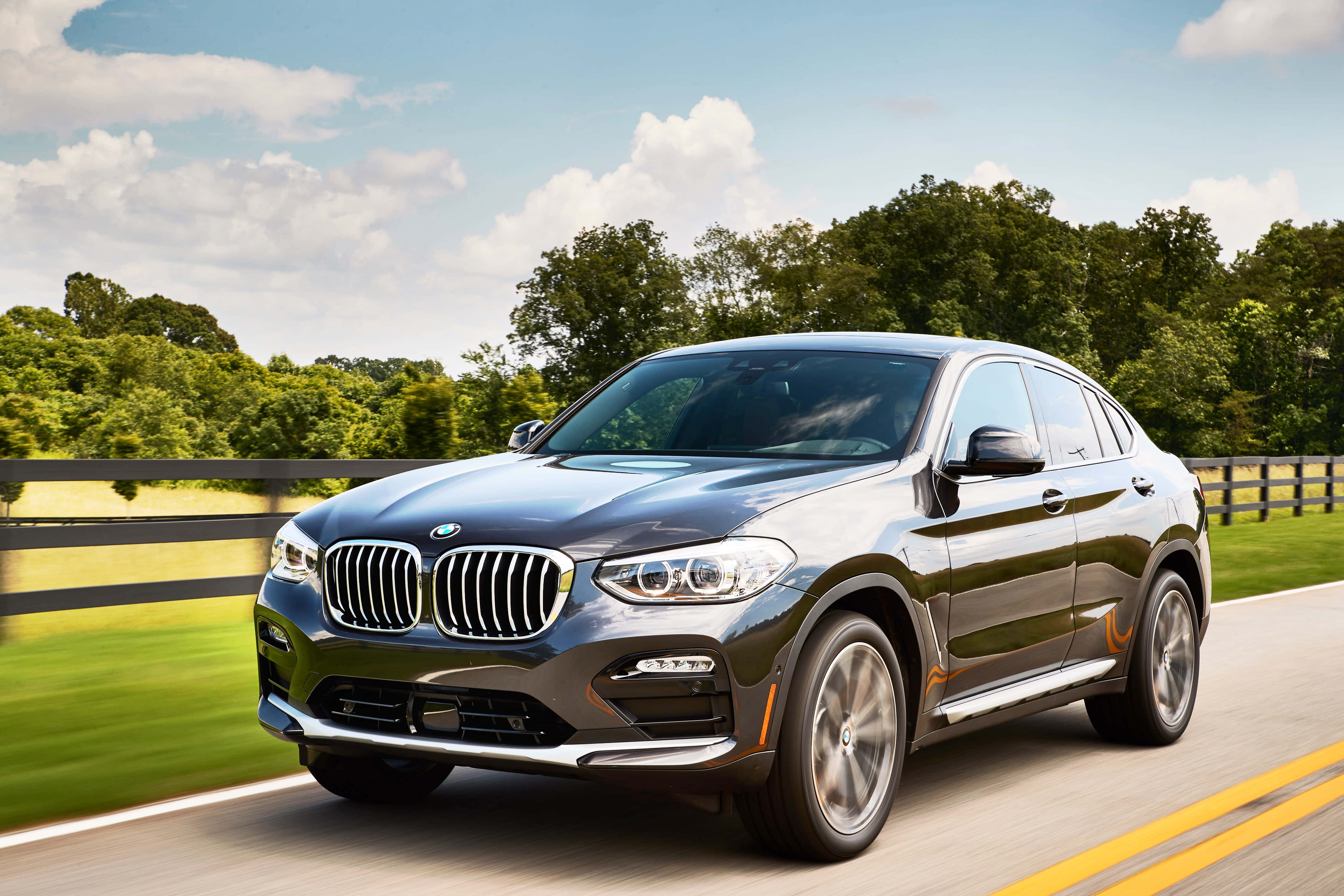 Bmw X4 桌布 壁紙 — 一輛時尚的灰色BMW X4在陽光明媚的鄉村道路上巡航，兩旁是茂密的綠樹和木柵欄。