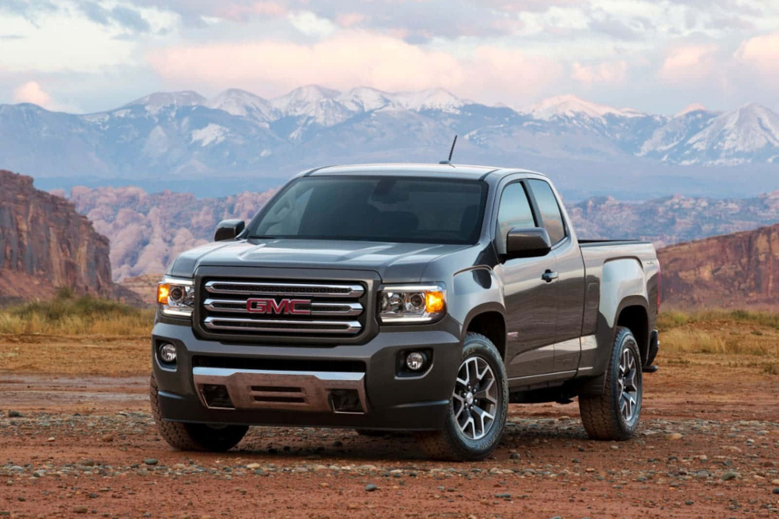 Gmc Canyon 桌布 壁紙 — 一輛時尚的灰色GMC Canyon皮卡車停在紅色沙漠泥土上，背景是白雪覆蓋的山脈和岩石峭壁，在柔和的暮色天空下。