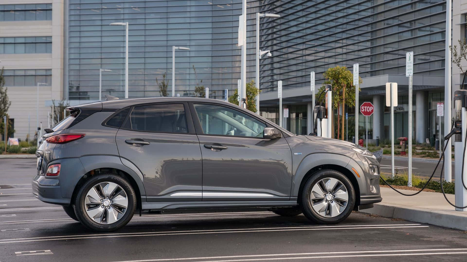 Hyundai Kona 桌布 壁紙 — 一輛時尚的灰色Hyundai Kona Electric在玻璃摩天大樓旁的現代城市充電站充電。