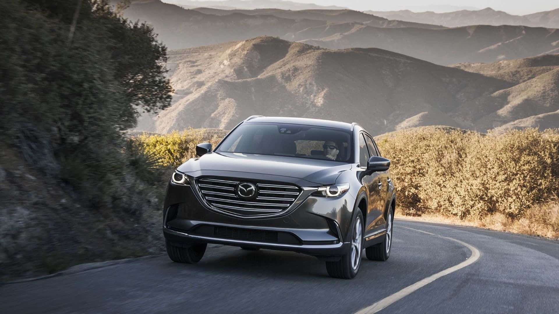 Mazda Cx-9 桌布 壁紙 — 一輛時尚的灰色Mazda CX-9 SUV在日出時沿著蜿蜒的山路穿過金色的山丘行駛。
