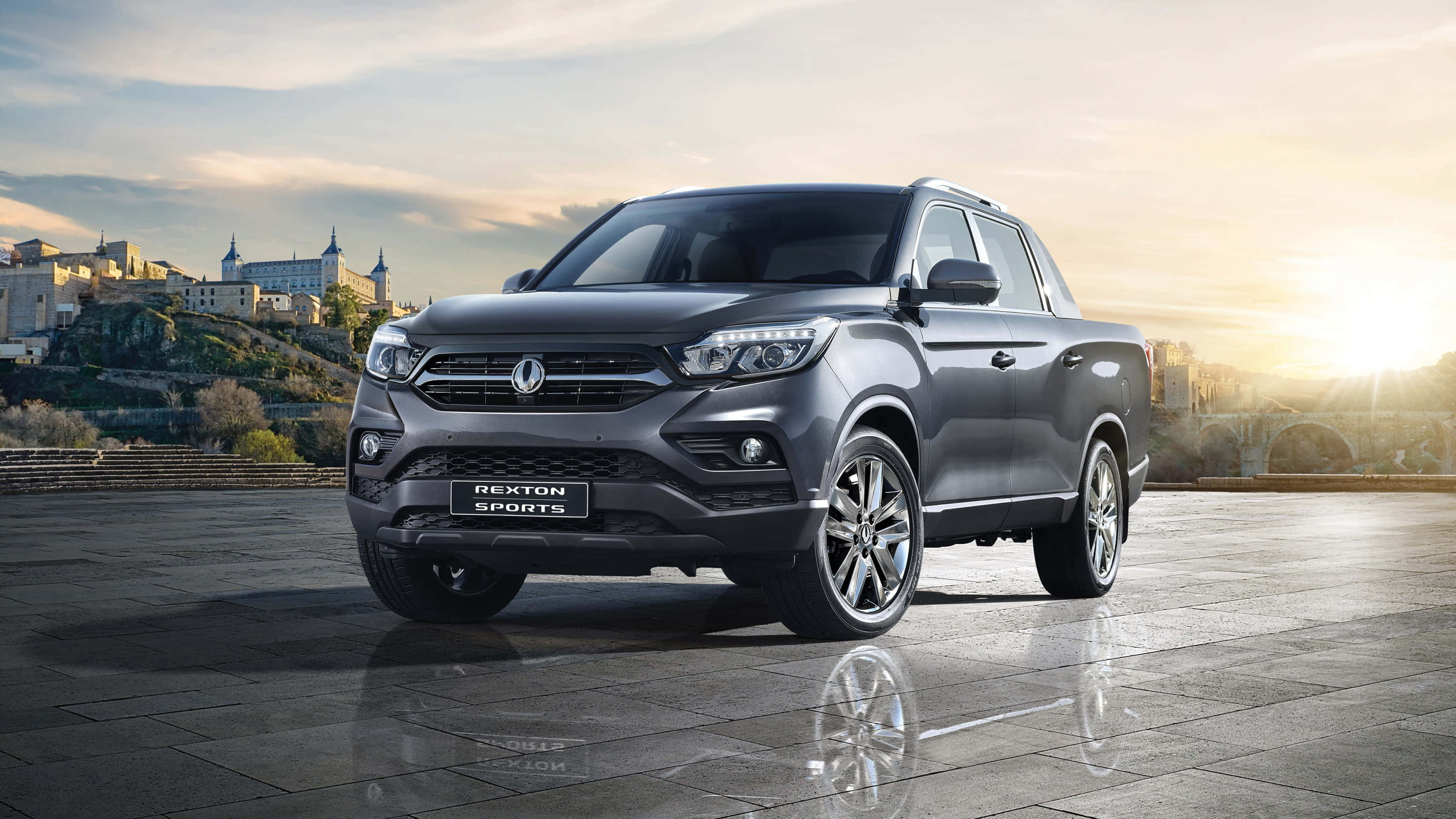 Ssangyong 桌布 壁紙 — 一輛時尚的灰色SsangYong Rexton Sports皮卡車在濕漉漉的石頭廣場上閃閃發光，金色的夕陽將歷史悠久的山城沐浴在溫暖的光芒中。