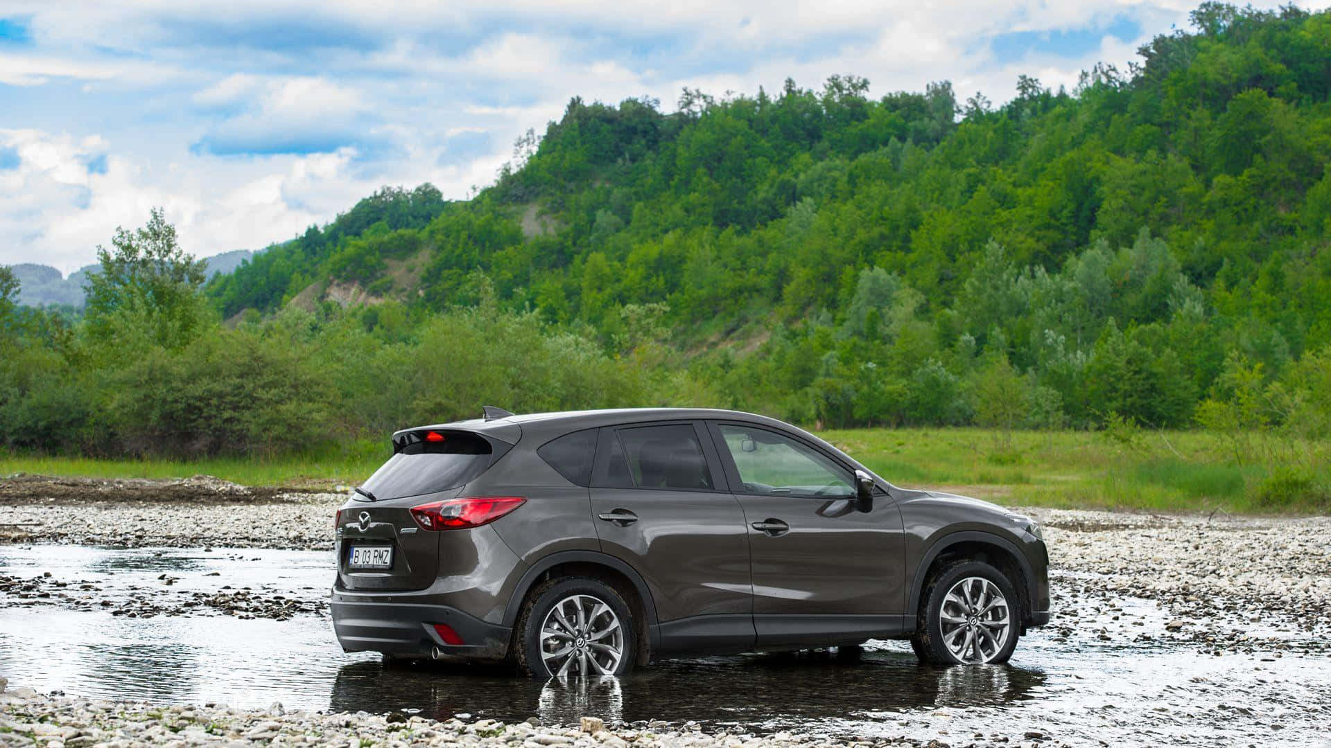 Mazda Cx-5 桌布 壁紙 — 一輛時尚的灰色SUV停在淺水中，旁邊是岩石河岸，背後是鬱鬱蔥蔥的綠色山丘，天空部分多雲。