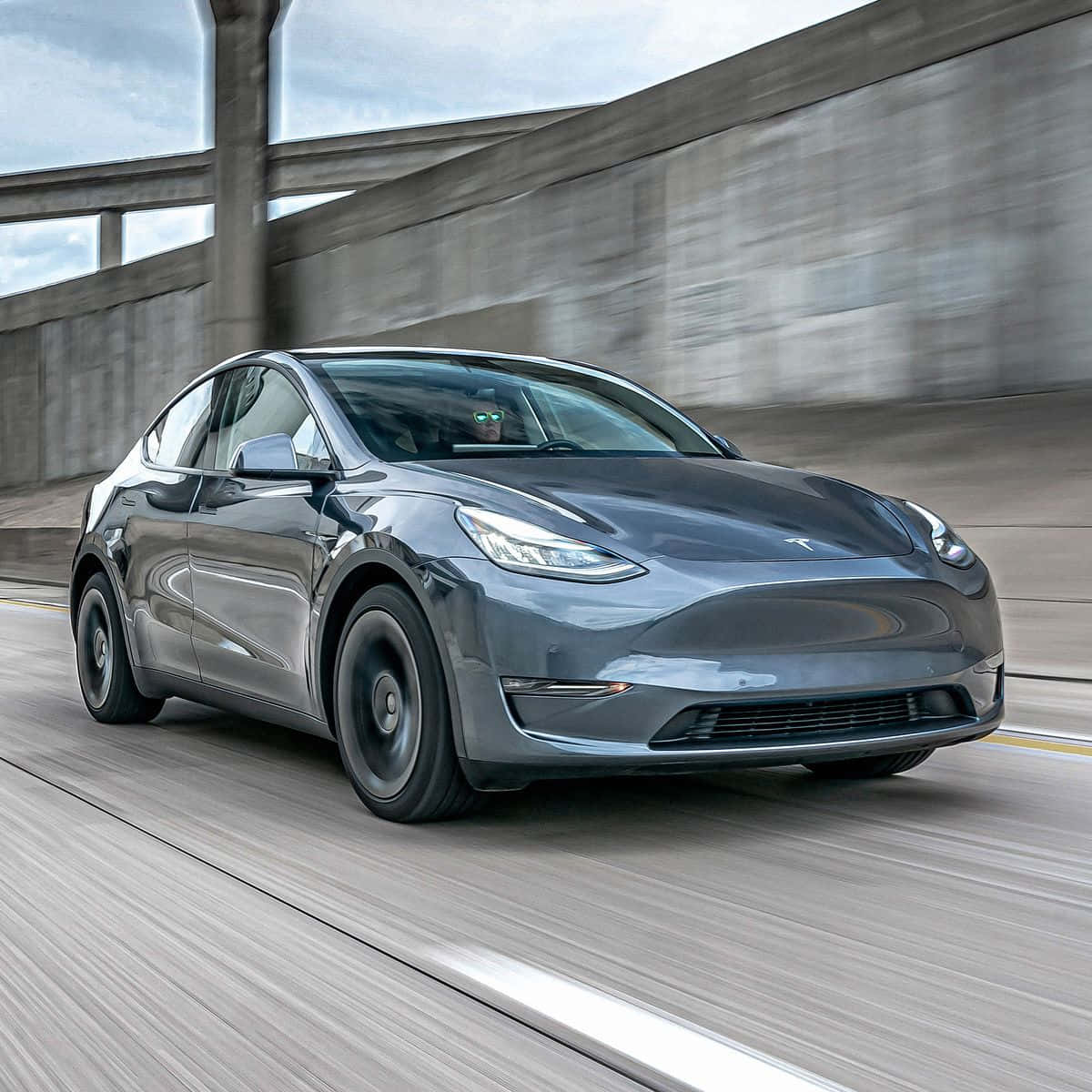 tesla 桌布 壁紙 — 一輛時尚的灰色Tesla Model Y在混凝土高速公路上行駛，經過天橋下方，動態模糊強調了速度。