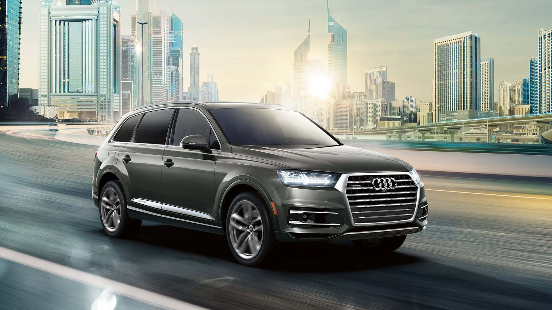 Audi Q7 桌布 壁紙 — 一輛時尚的灰色奧迪SUV在高速公路上飛馳，背後是現代城市天際線在金色夕陽中閃耀。