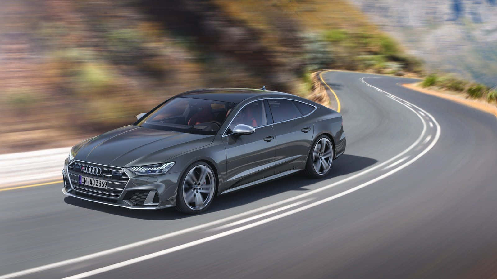 Audi S7 桌布 壁紙 — 一輛時尚的灰色奧迪房車沿著蜿蜒的山路疾馳，背景是模糊的樹葉和遠處的山峰。