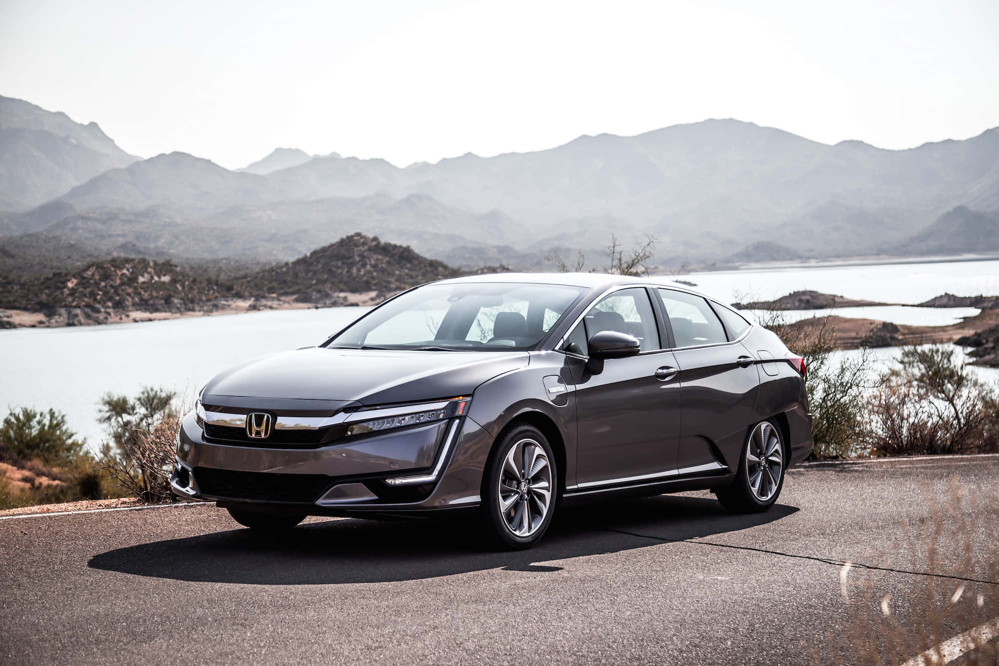 Honda Clarity 桌布 壁紙 — 一輛時尚的灰色本田Clarity插電式混合動力車停在寧靜湖泊旁的鋪砌道路上，遠山在明亮的天空下。