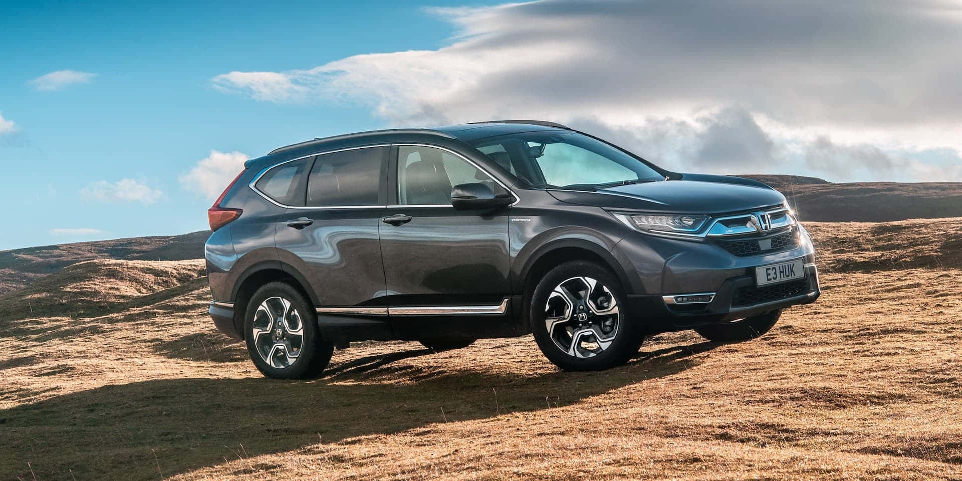Honda Cr-v 桌布 壁紙 — 一輛時尚的灰色本田CR-V SUV停放在乾草地上，背景是部分多雲的藍天和起伏的丘陵。