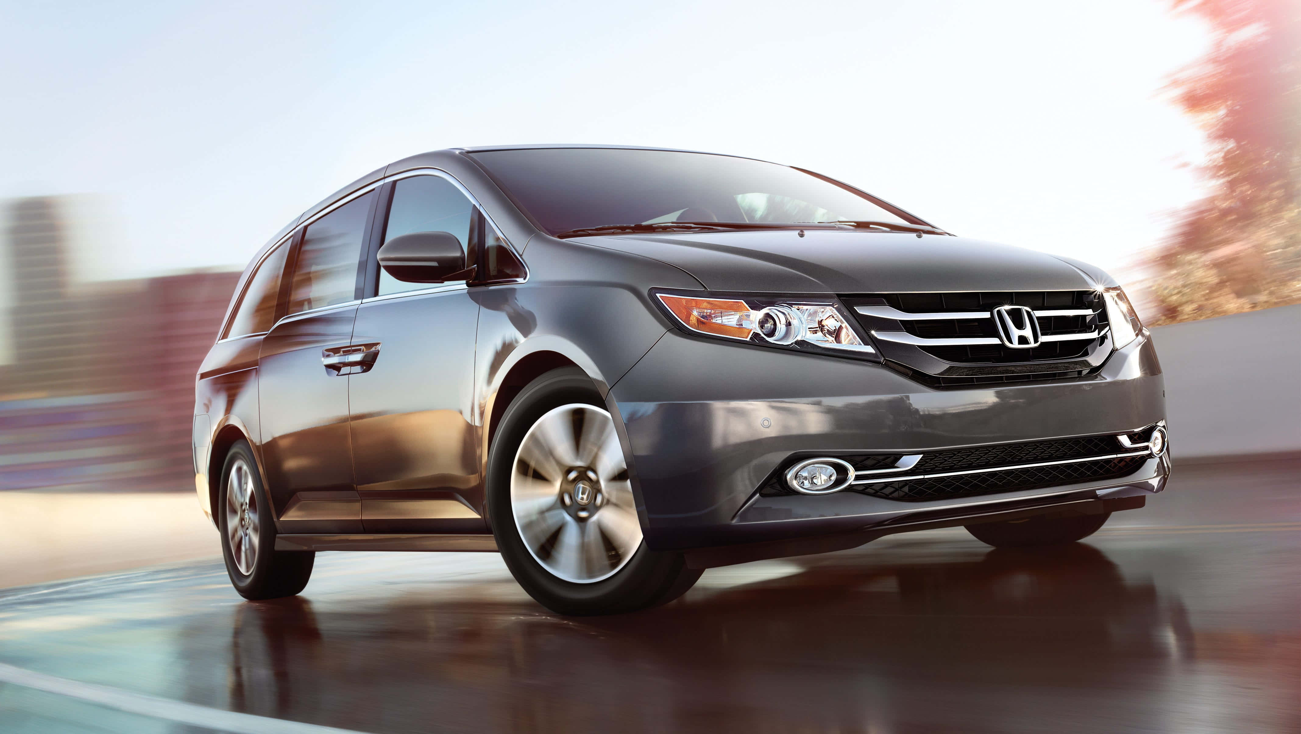 Honda Odyssey 桌布 壁紙 — 一輛時尚的灰色本田小型貨車在潮濕的道路上行駛，背景是模糊的城市景觀和溫暖的日落餘暉。