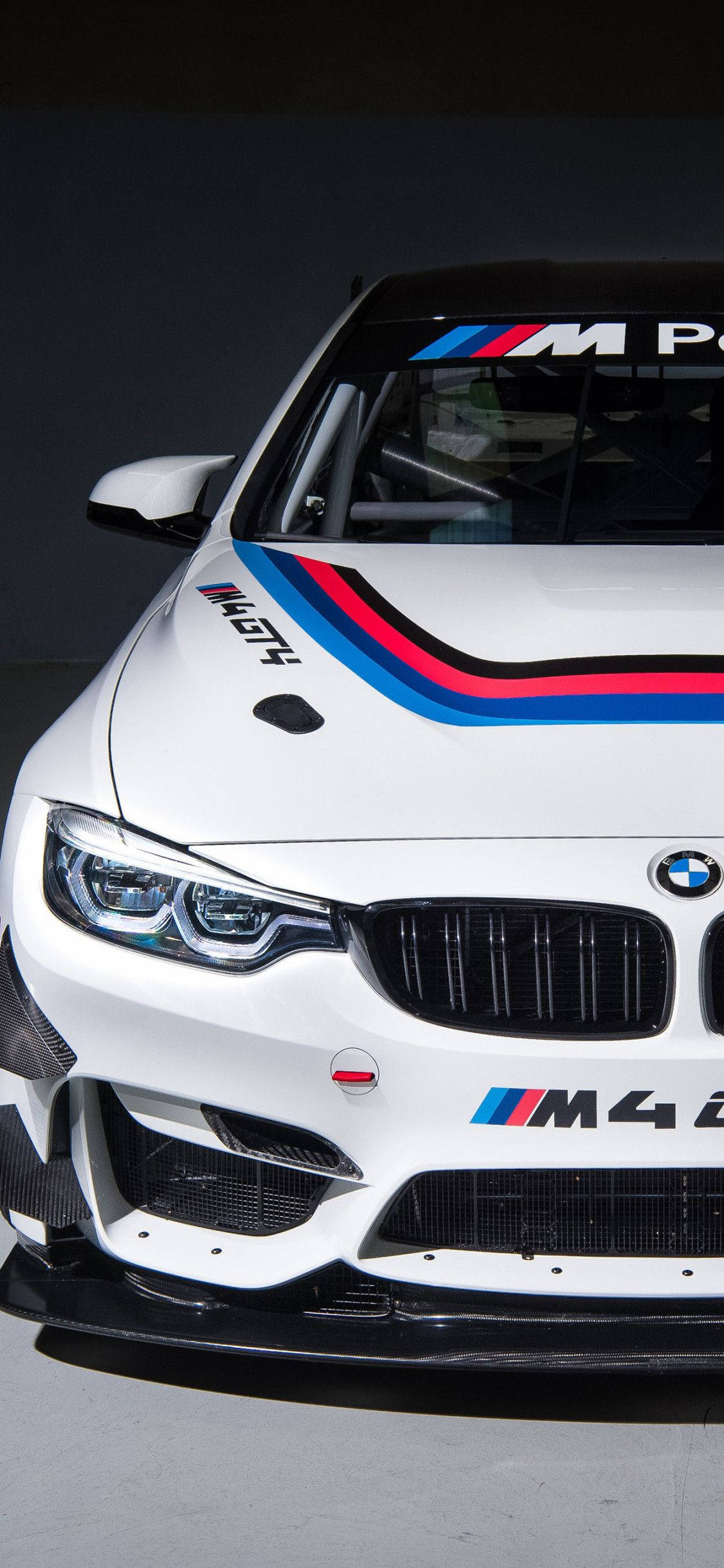 M4 GT4 BMW iPhone X 壁紙 — 一輛時尚的白色BMW M4賽車，帶有醒目的藍、紅、黑條紋，在戲劇性的攝影棚拍攝中捕捉到其侵略性的前臉和賽車貼花。