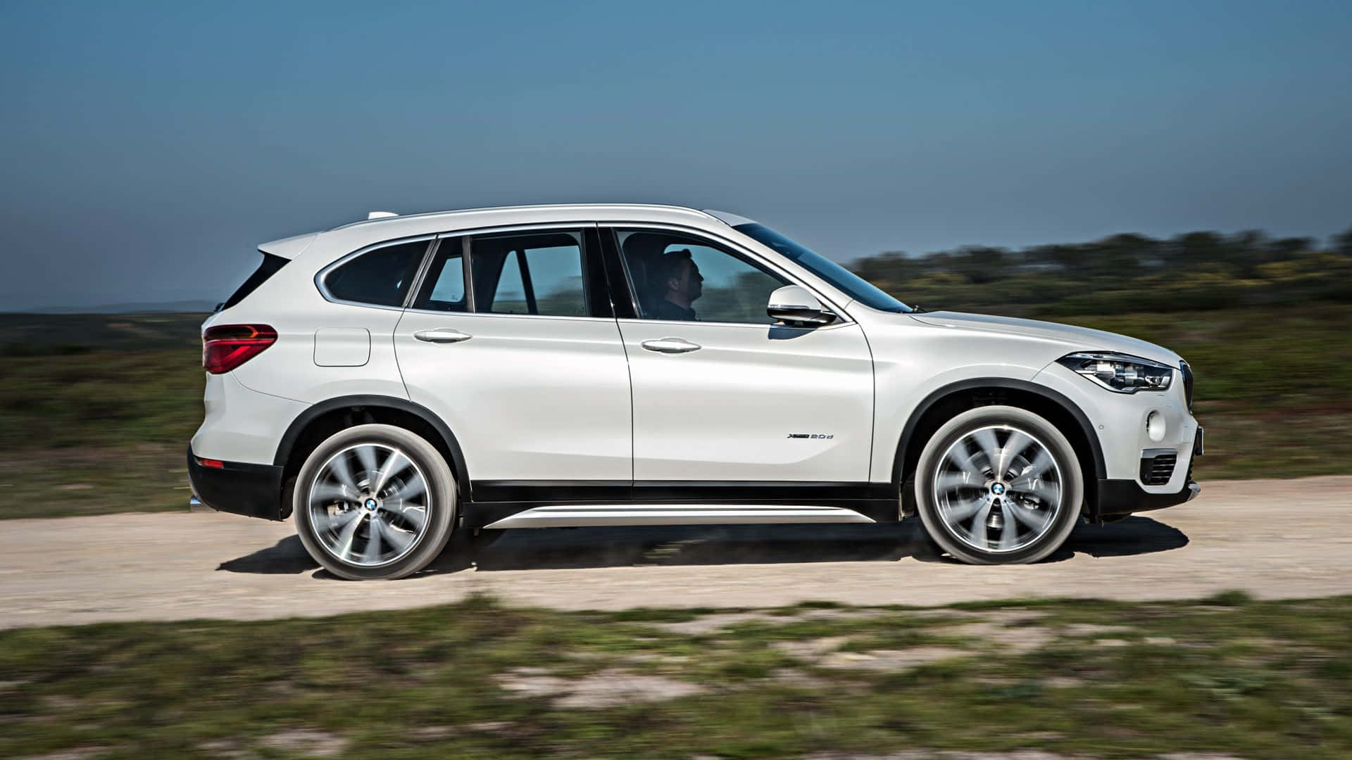 Bmw X1 桌布 壁紙 — 一輛時尚的白色BMW SUV在陽光明媚的泥路上行駛，背景是模糊的綠意和藍天。