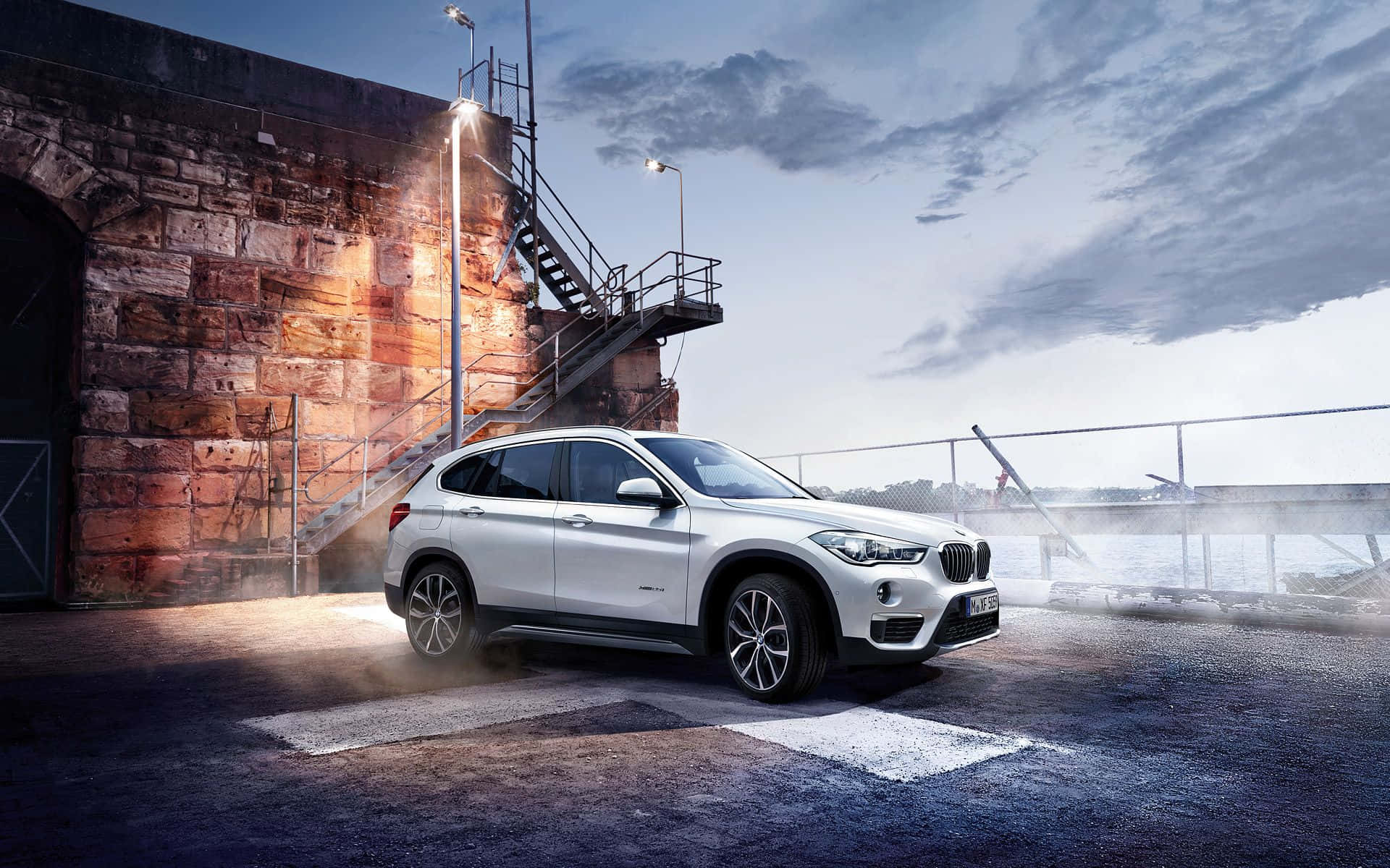 Bmw X1 桌布 壁紙 — 一輛時尚的白色BMW SUV在黃昏時停泊在石倉庫旁，帶有戲劇性的燈光和霧氣瀰漫的氛圍。