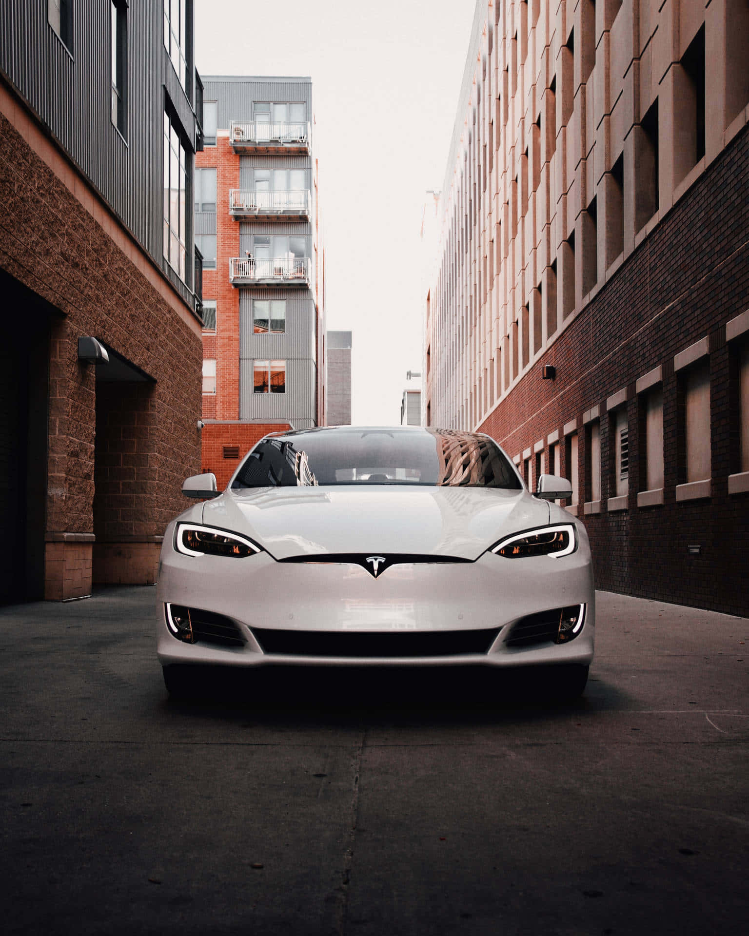 tesla 桌布 壁紙 — 一輛時尚的白色Tesla Model S在狹窄的城市小巷中向前，兩旁是磚砌和現代建築，天空多雲。