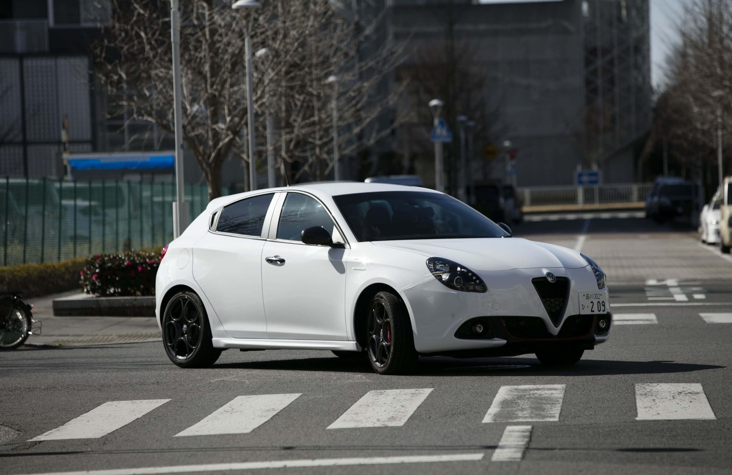 Alfa Romeo Giulietta 桌布 壁紙 — 一輛時尚的白色愛快羅密歐Giulietta配黑色輪圈，在陽光普照的市區街道上行駛，街道上有行人過路處。