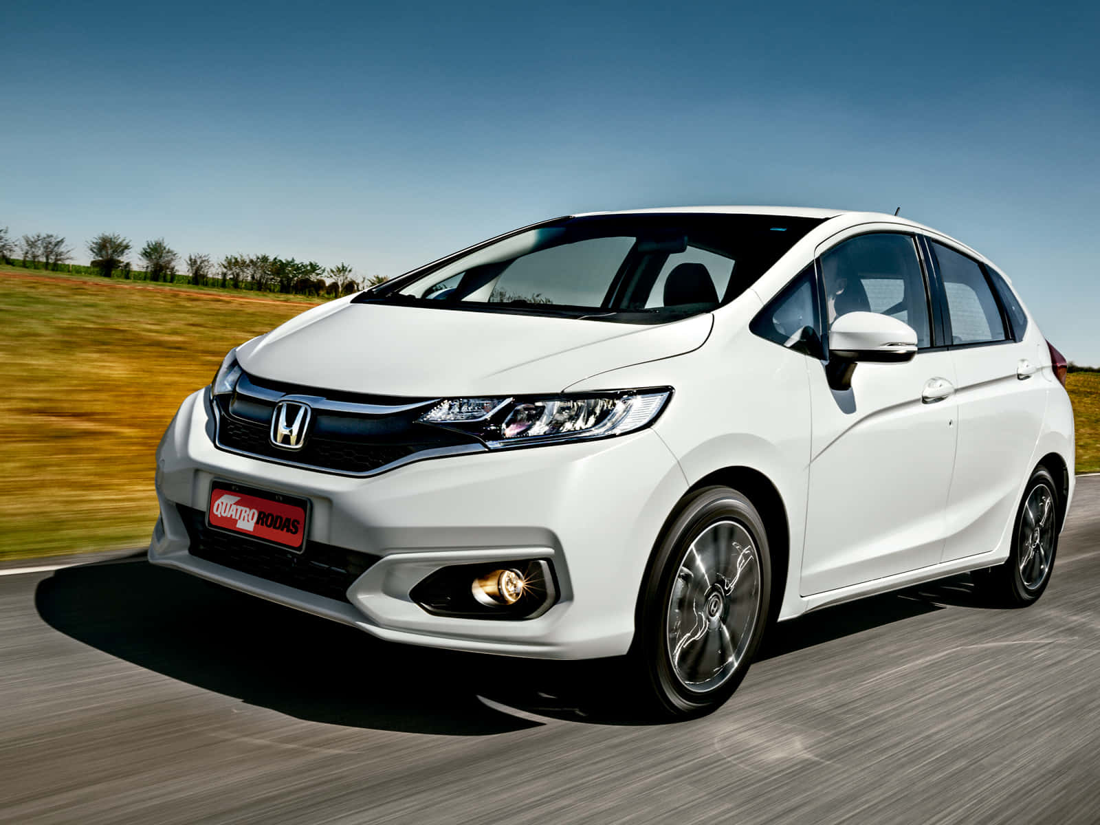 Honda Fit 桌布 壁紙 — 一輛時尚的白色本田Fit在陽光明媚的道路上行駛，背景是模糊的金色田野和藍天。