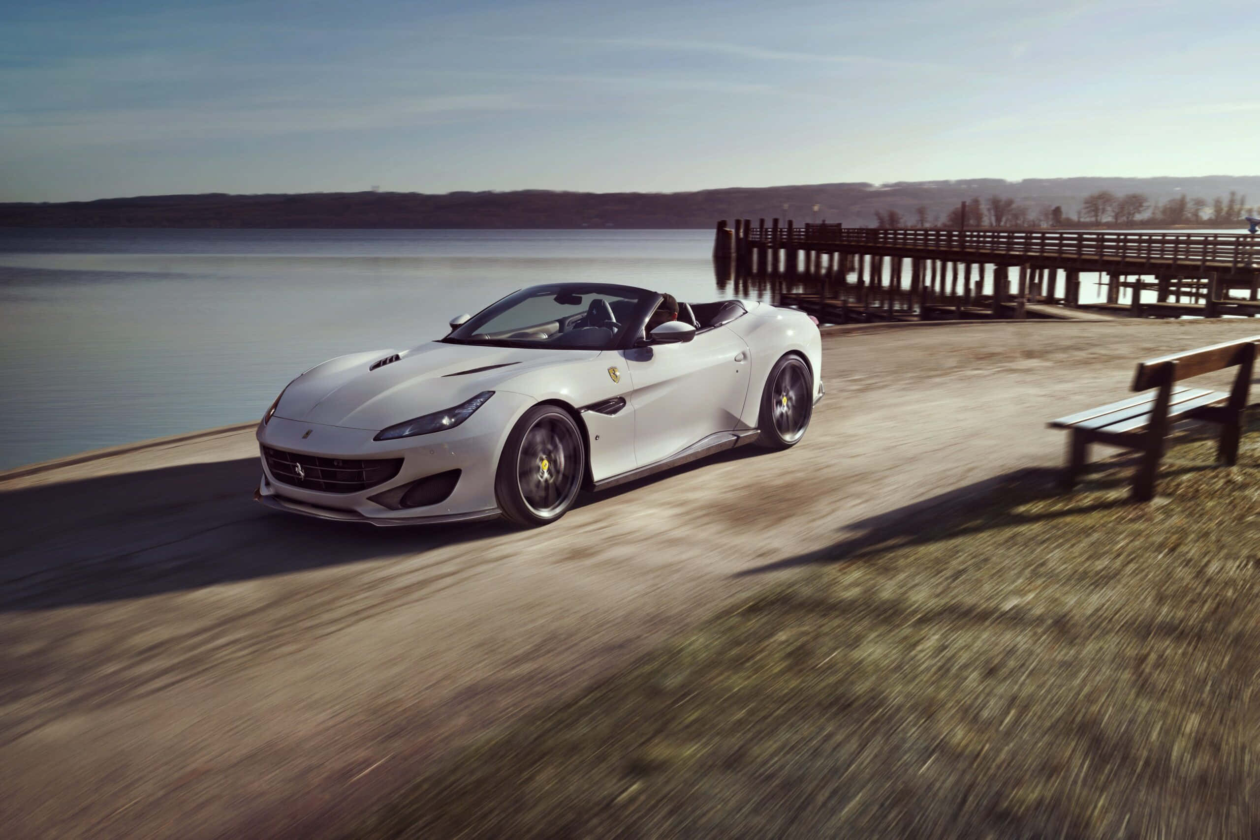 Ferrari Portofino 桌布 壁紙 — 一輛時尚的白色法拉利Portofino敞篷車在明亮的日光下沿著湖邊小路疾馳，帶有動態模糊效果。