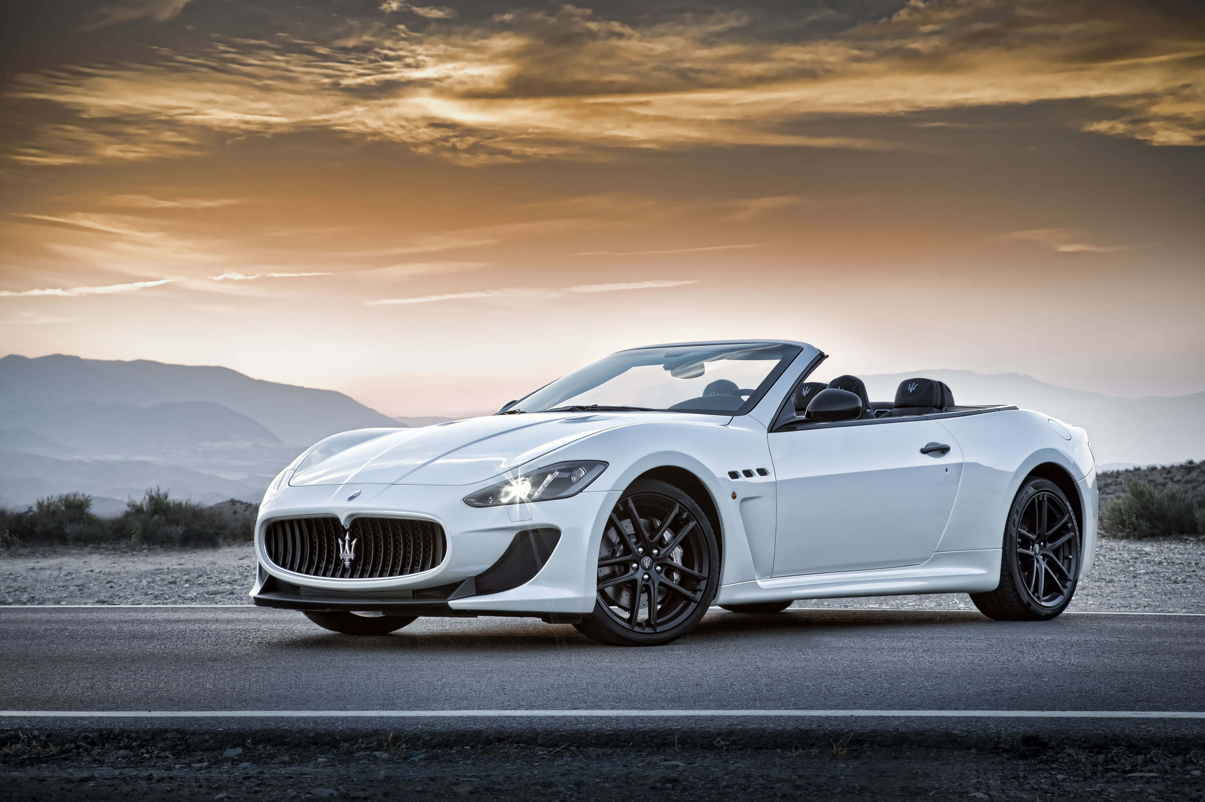 White Maserati 4K 壁紙 — 一輛時尚的白色瑪莎拉蒂敞篷車在金色日落天空和遠山下的沙漠公路上閃閃發光。