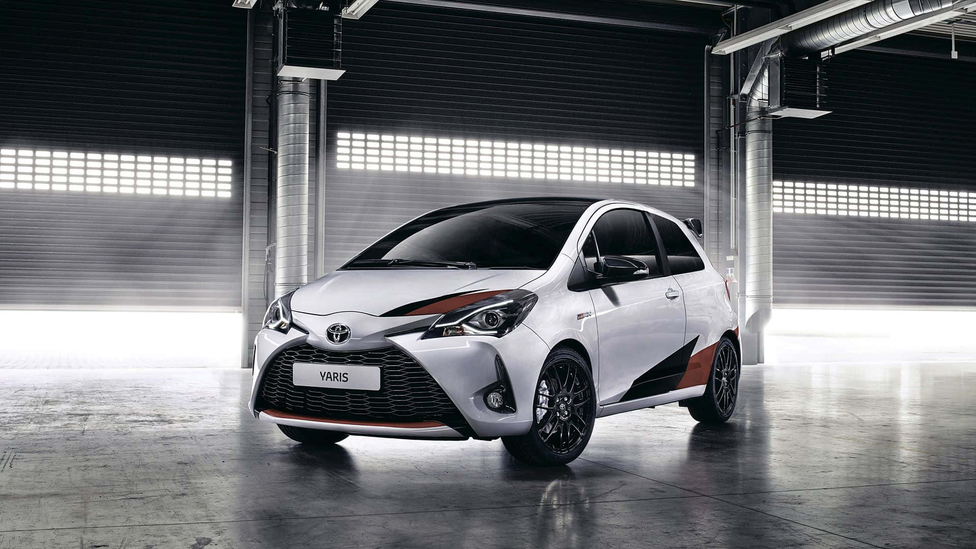 Toyota 桌布 壁紙 — 一輛時尚的白色豐田Yaris帶有橙色點綴，在明亮的頂燈下於現代工業車庫中展示。