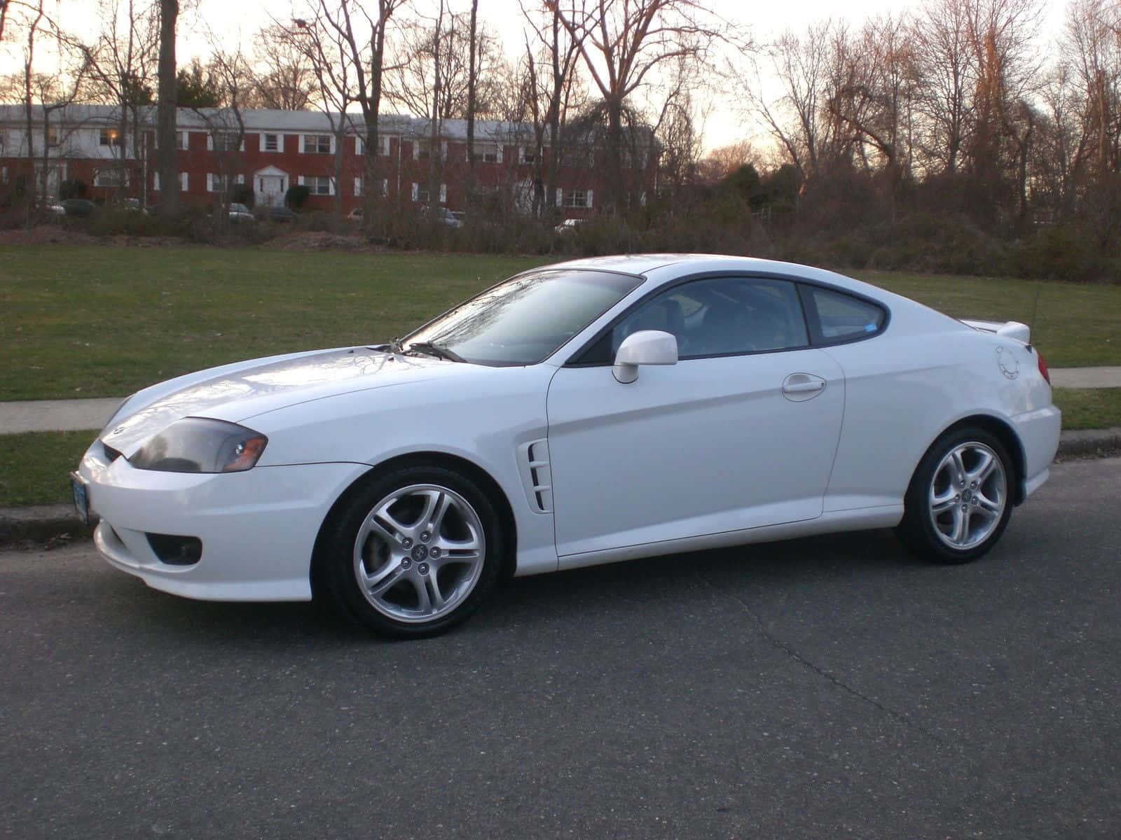 Hyundai Tiburon 桌布 壁紙 — 一輛時尚的白色運動轎跑車停在日落時分的街道上，背景是光禿禿的樹木和磚砌建築。