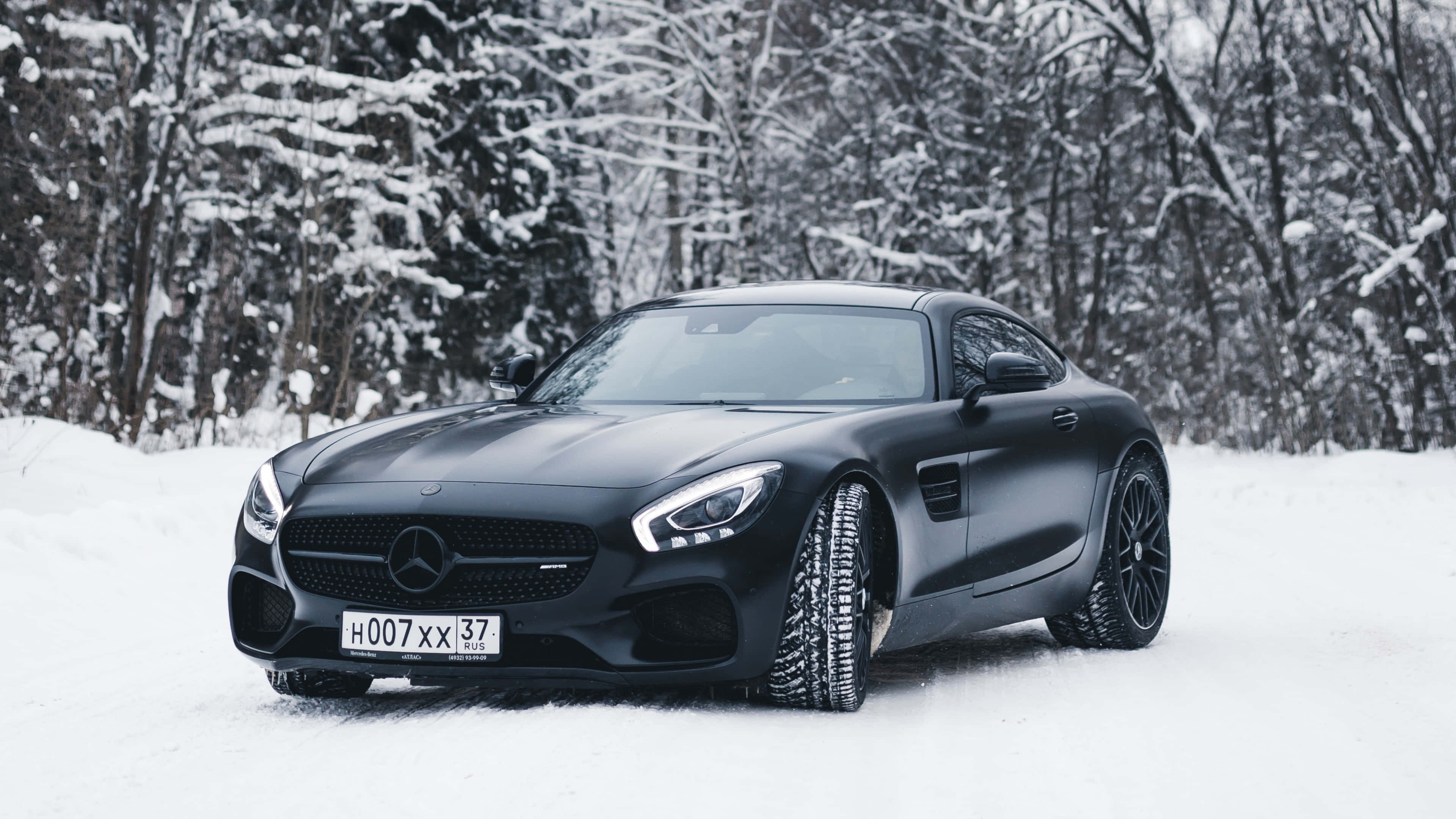 4K Mercedes 桌布 壁紙 — 一輛時尚的磨砂黑色Mercedes-AMG GT跑車停在積雪的森林道路上，背景是白雪覆蓋的樹木。