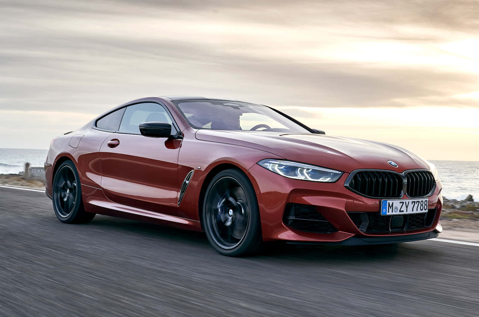 Bmw 8 Series 桌布 壁紙 — 一輛時尚的紅色BMW雙門跑車在日落時分沿著海岸公路飛馳，背景是海浪。