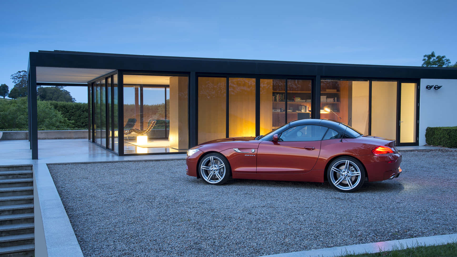 Bmw Z4 桌布 壁紙 — 一輛時尚的紅色跑車停泊在碎石路上，前方是一座在黃昏時分溫暖發光的現代玻璃屋。
