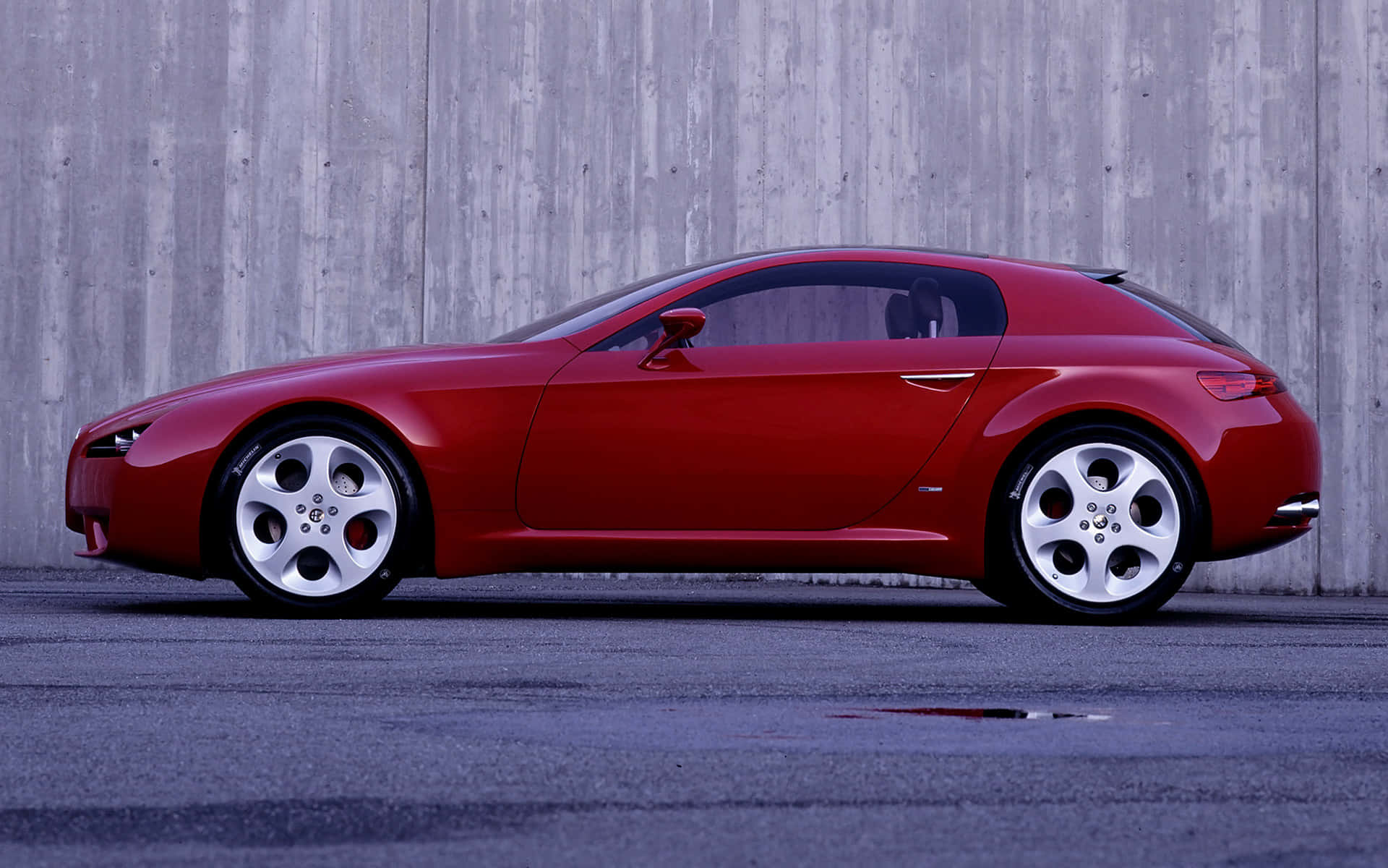 Alfa Romeo Brera 桌布 壁紙 — 一輛時尚的紅色跑車配備銀色輪圈，停泊在風化的灰色混凝土牆旁，地面為鋪砌表面。