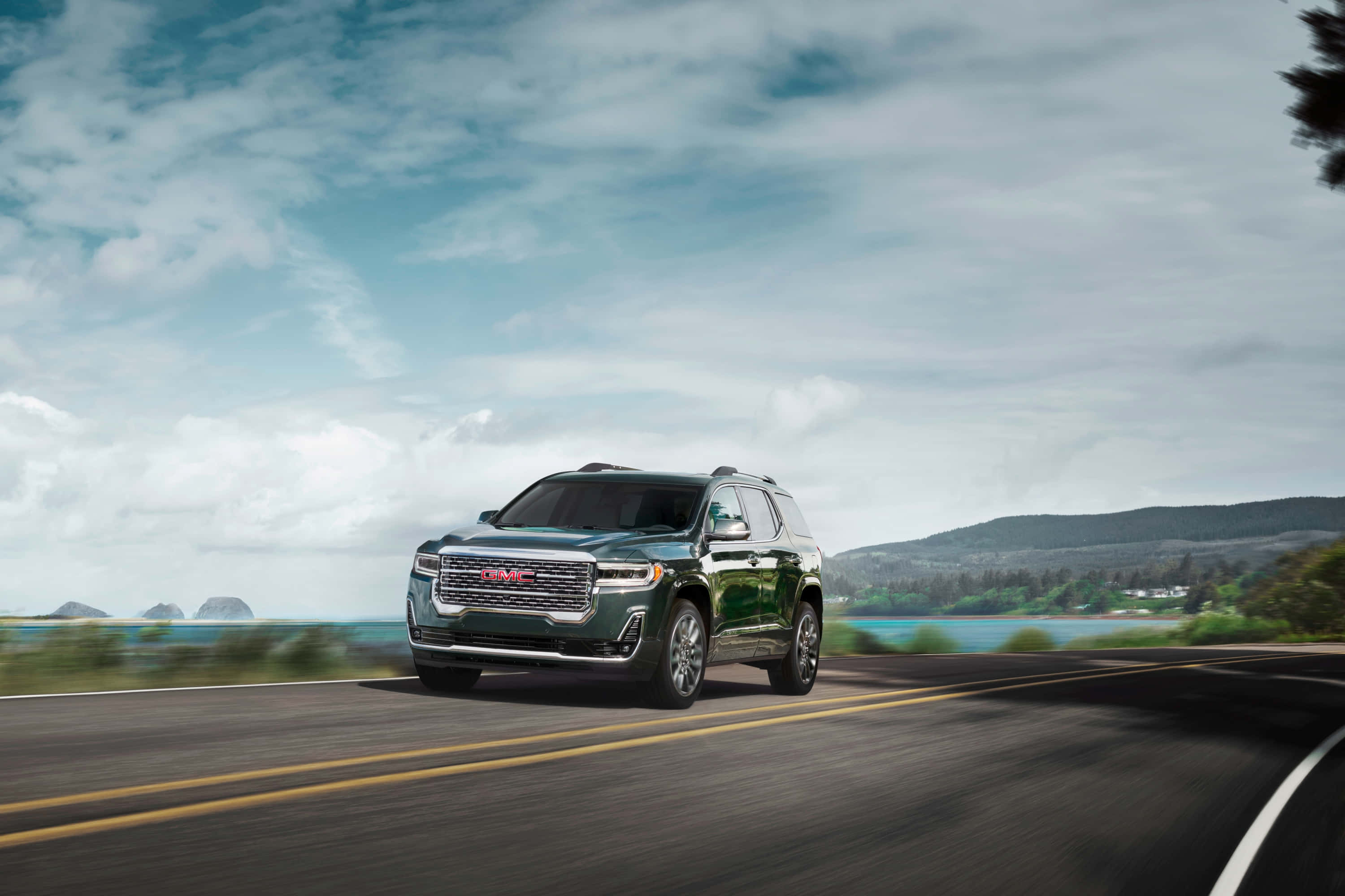 Gmc Acadia 桌布 壁紙 — 一輛時尚的綠色GMC SUV在部分多雲的天空下，沿著海岸高速公路行駛，帶有模糊的動態效果。