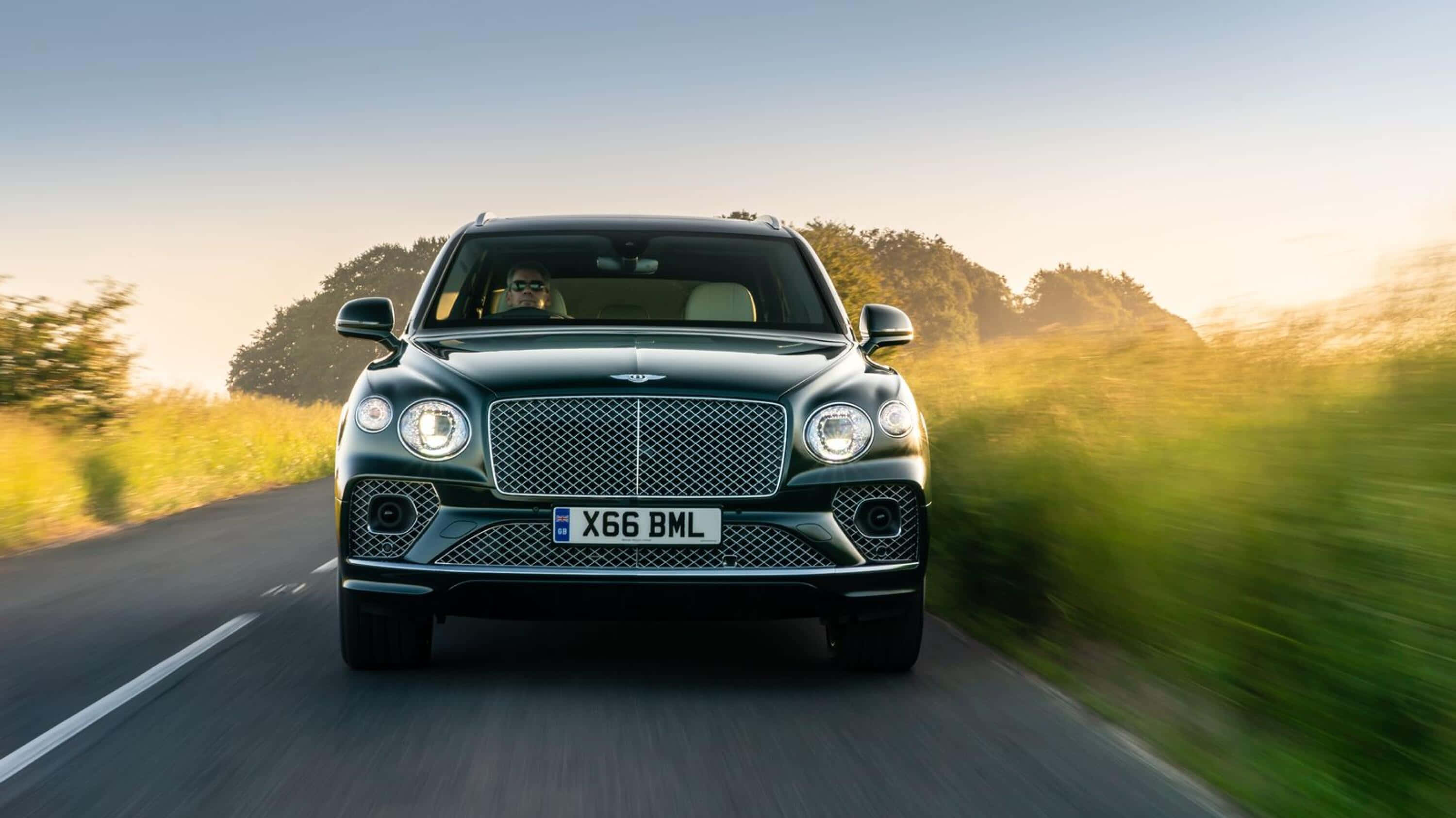 Bentley Bentayga 桌布 壁紙 — 一輛時尚的綠色賓利 Bentayga SUV 在陽光明媚的鄉村道路上飛馳，背景是模糊的金色草地和樹木。