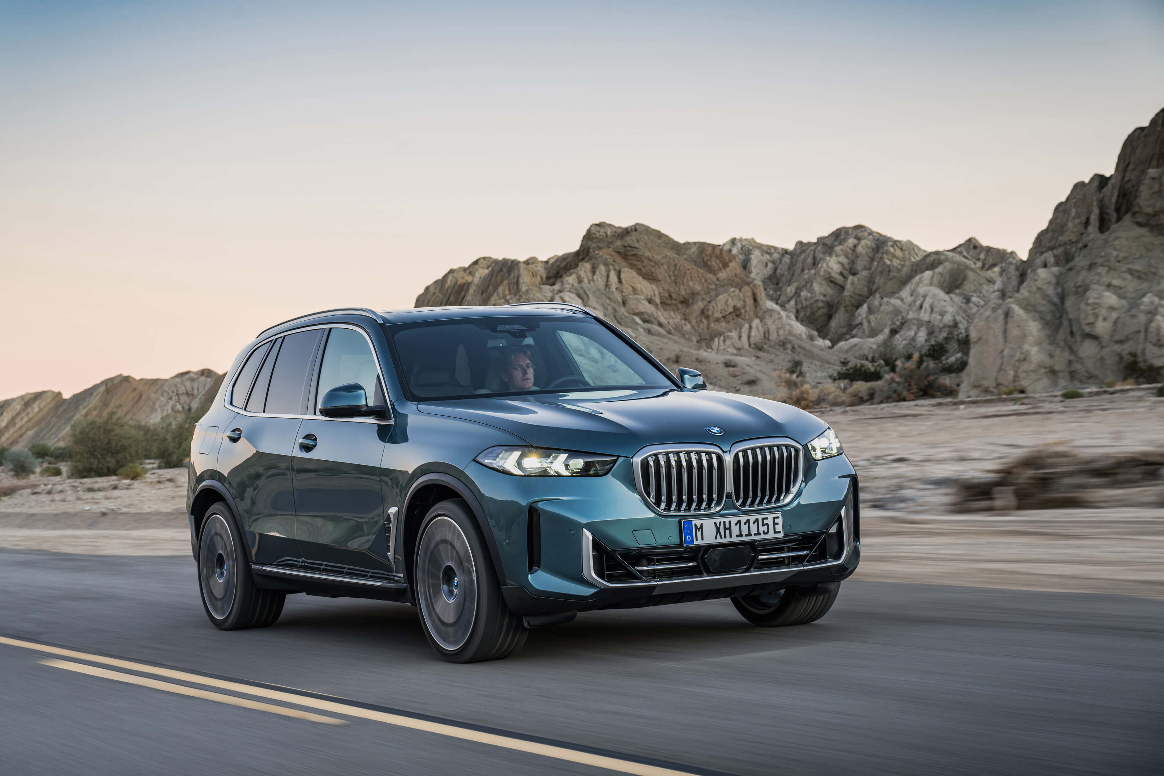 Bmw X5 桌布 壁紙 — 一輛時尚的藍綠色BMW X5 SUV在柔和的暮色天空下，沿著一條有崎嶇岩石地貌的沙漠道路行駛。