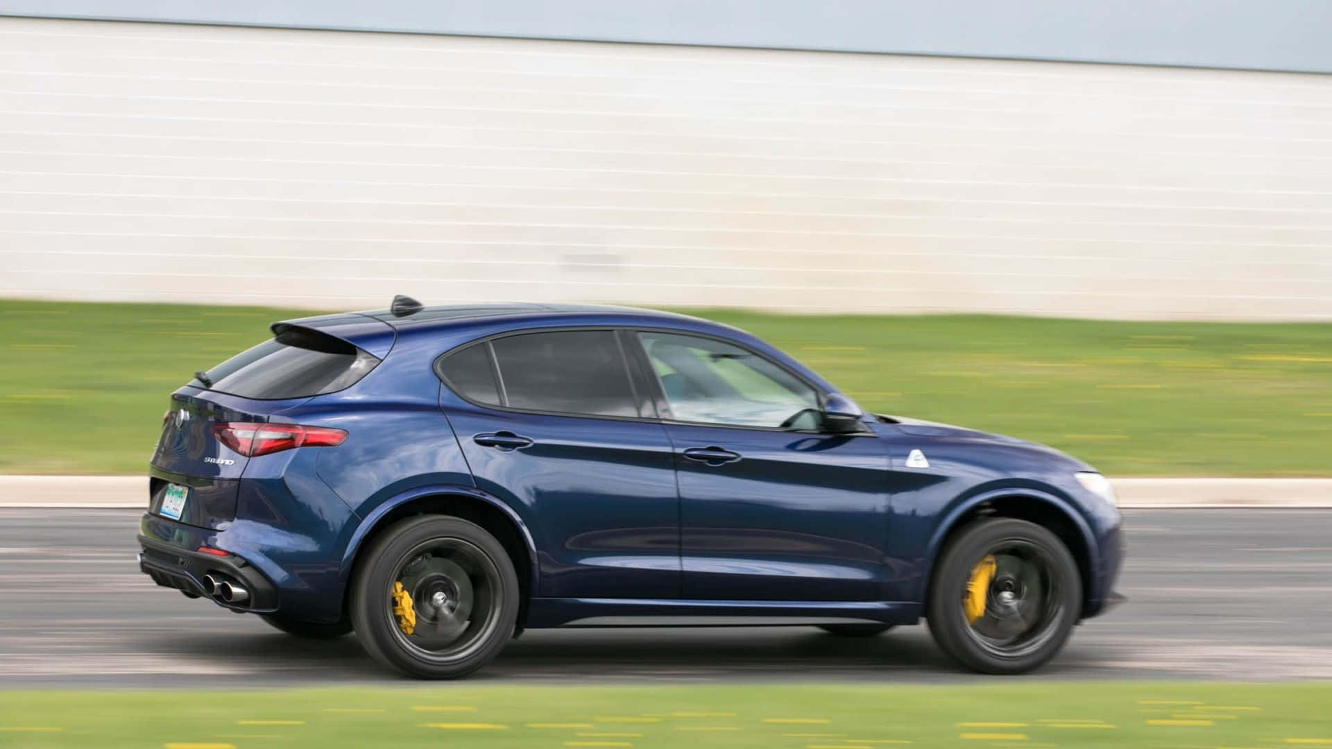 Alfa Romeo Stelvio 桌布 壁紙 — 一輛時尚的藍色 Alfa Romeo Stelvio SUV 在賽道上疾馳，背景是模糊的綠色草地和白色牆壁。