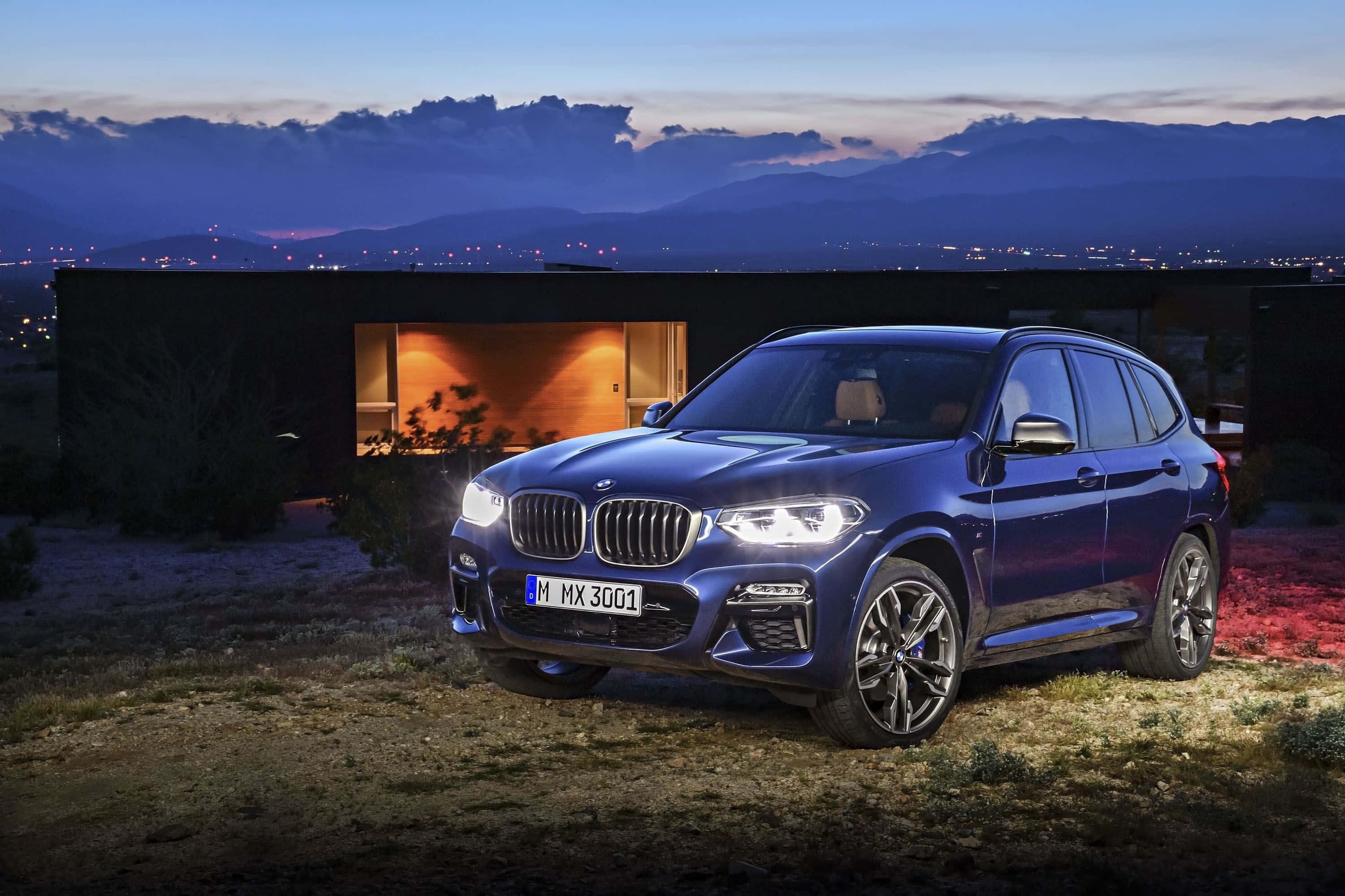 Bmw X3 桌布 壁紙 — 一輛時尚的藍色BMW X3 SUV在黃昏時停在一棟現代房屋前，溫暖的燈光在暮色山巒背景下閃耀。