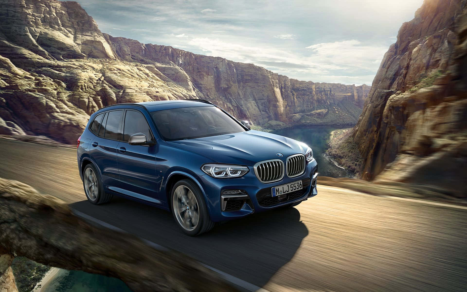 Bmw X3 桌布 壁紙 — 一輛時尚的藍色BMW X3 SUV在明亮的天空下，沿著蜿蜒的峽谷道路疾馳，兩旁是崎嶇的懸崖和遠處的河流。