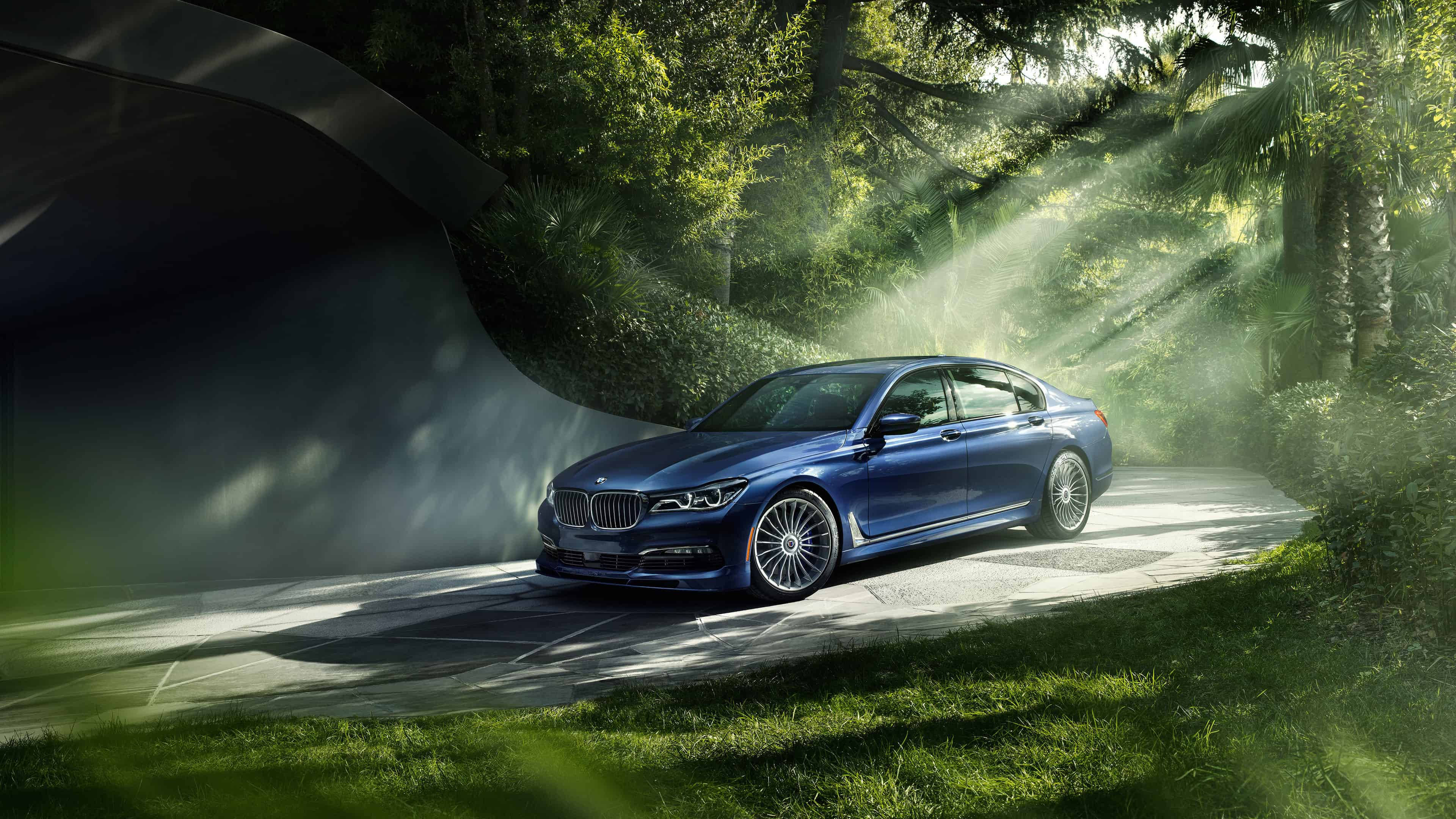 4K BMW Alpina B7 壁紙 — 一輛時尚的藍色BMW轎車沿著一條霧氣瀰漫的森林道路行駛，沐浴在透過茂密綠樹灑落的金色陽光中。
