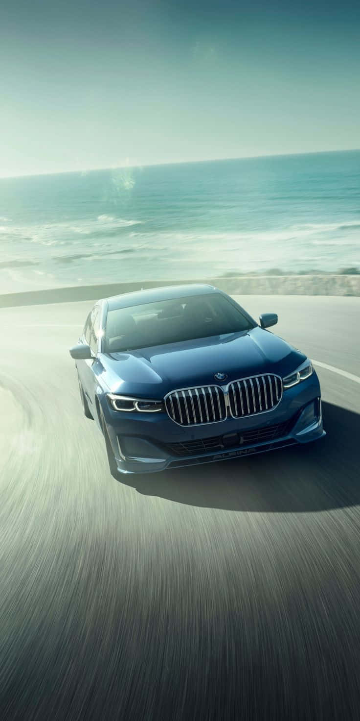 Bmw 7 Series 桌布 壁紙 — 一輛時尚的藍色BMW轎車沿著海岸公路飛馳，背景是晴朗天空下拍打的海浪。