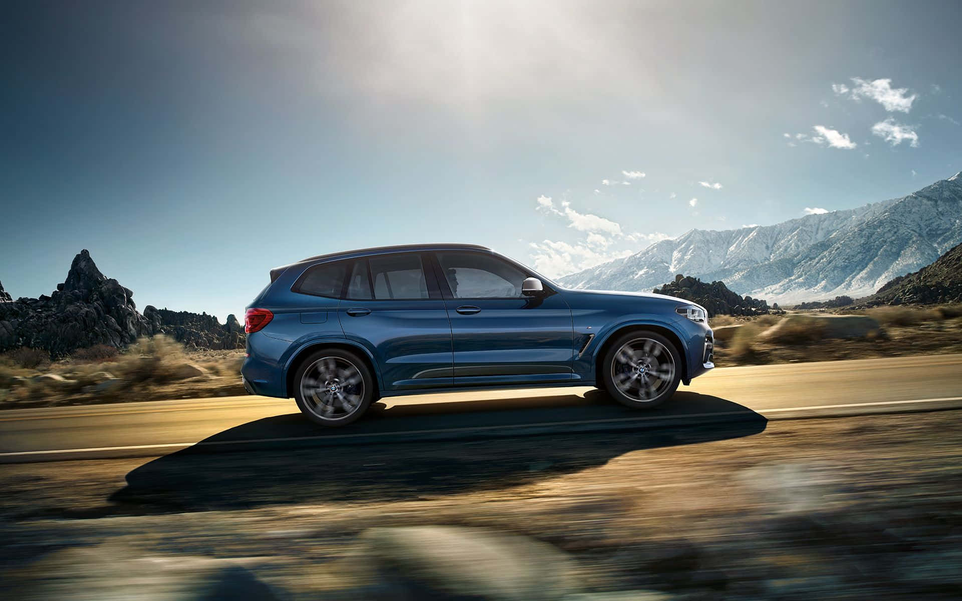 Bmw X3 桌布 壁紙 — 一輛時尚的藍色SUV在烈日下沿著沙漠公路行駛，兩旁是崎嶇的山脈和岩石露頭。