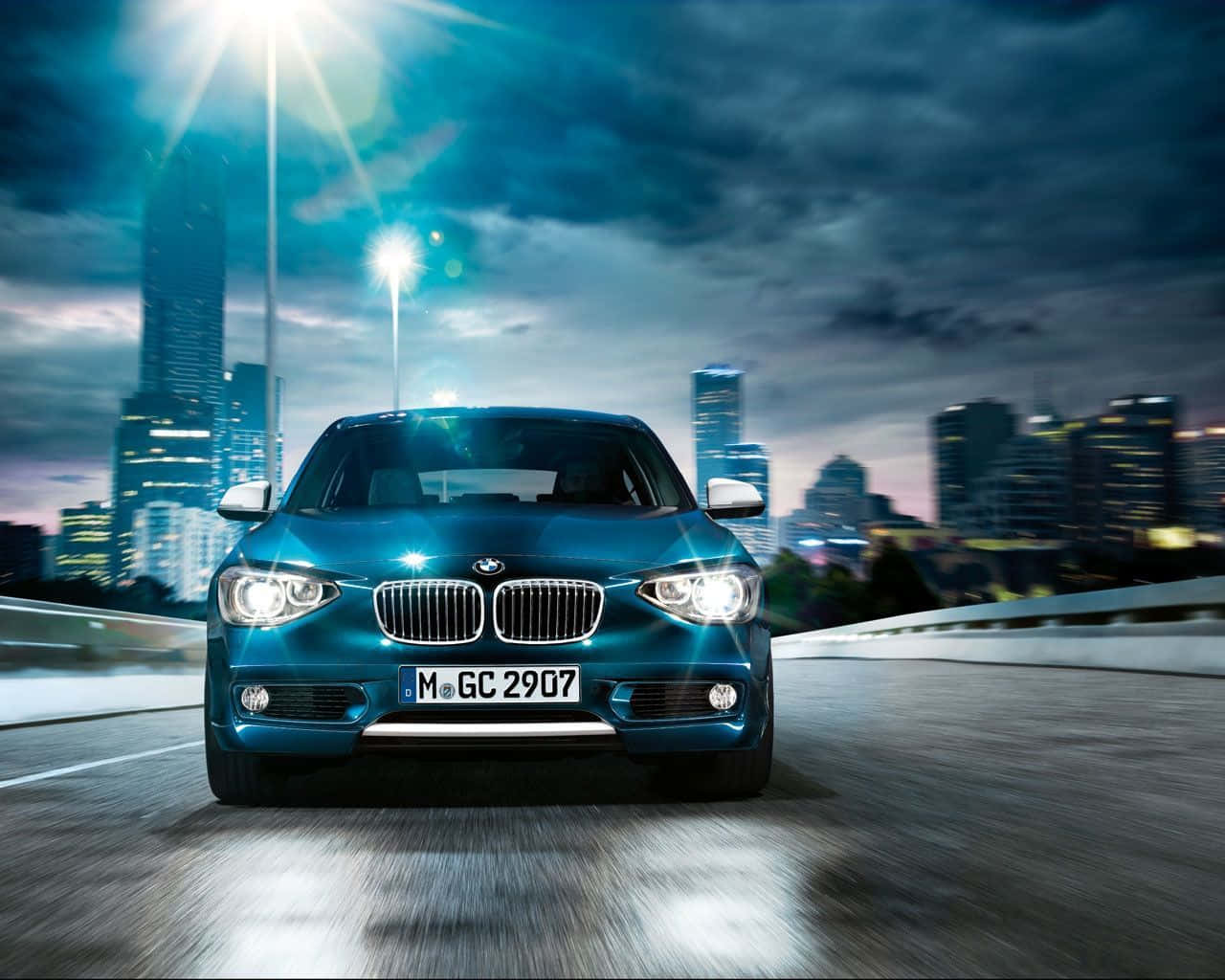 Bmw 1 Series 桌布 壁紙 — 一輛時尚的藍色寶馬轎車在濕漉漉的城市橋樑上，在戲劇性的暮色天空下，閃爍的路燈和摩天大樓中向觀眾疾馳。