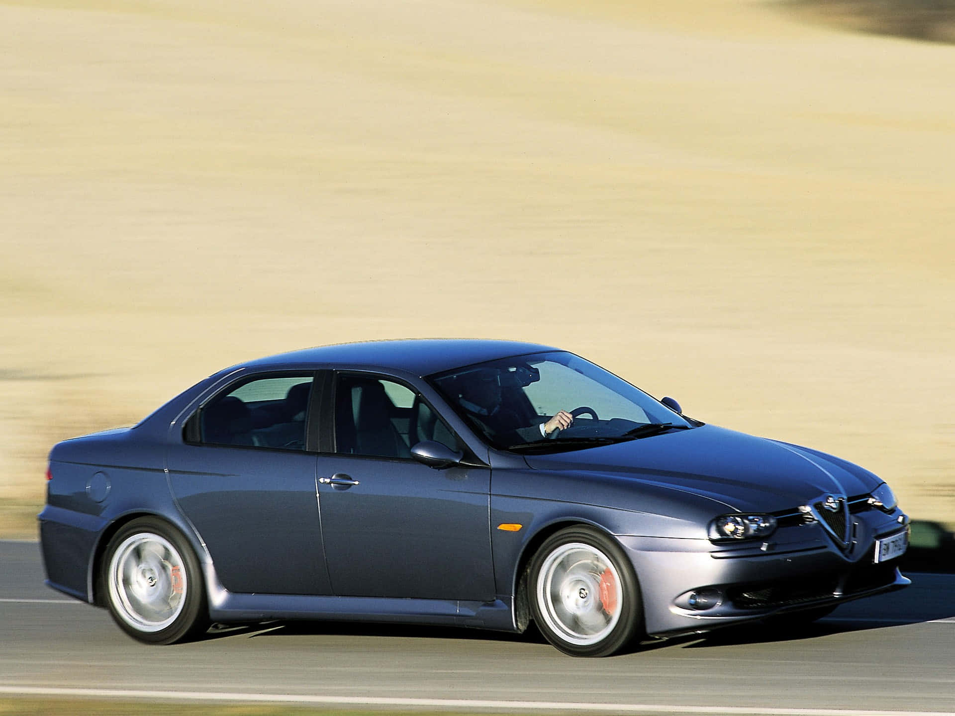 Alfa Romeo 156 桌布 壁紙 — 一輛時尚的藍色愛快羅密歐轎車在陽光明媚的道路上行駛，背景是模糊的金色田野。