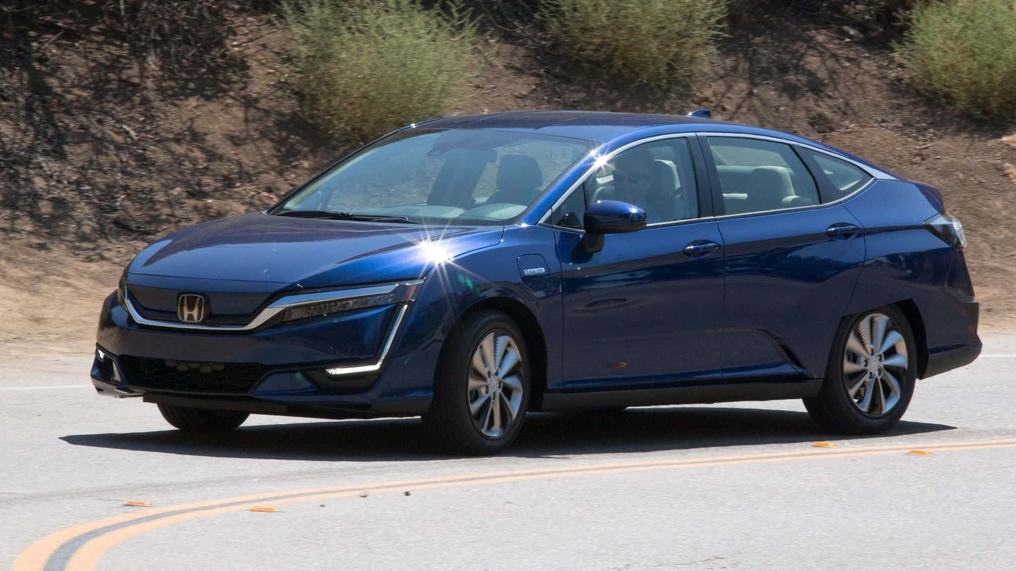 Honda Clarity 桌布 壁紙 — 一輛時尚的藍色本田Clarity插電式混合動力轎車行駛在陽光明媚的沙漠公路上，兩旁是乾燥的灌木山坡。