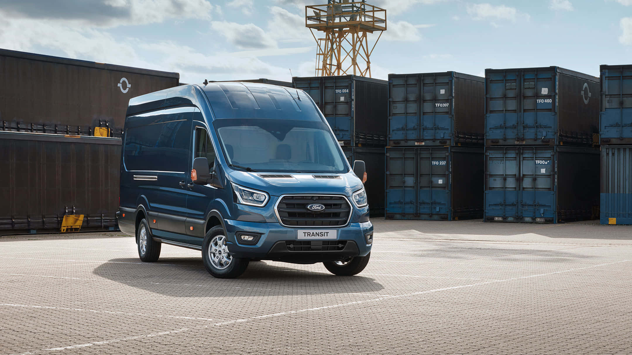 Ford Transit 桌布 壁紙 — 一輛時尚的藍色福特 Transit 貨車停泊在鋪砌的庭院中，周圍堆疊著貨運集裝箱，天空部分多雲。