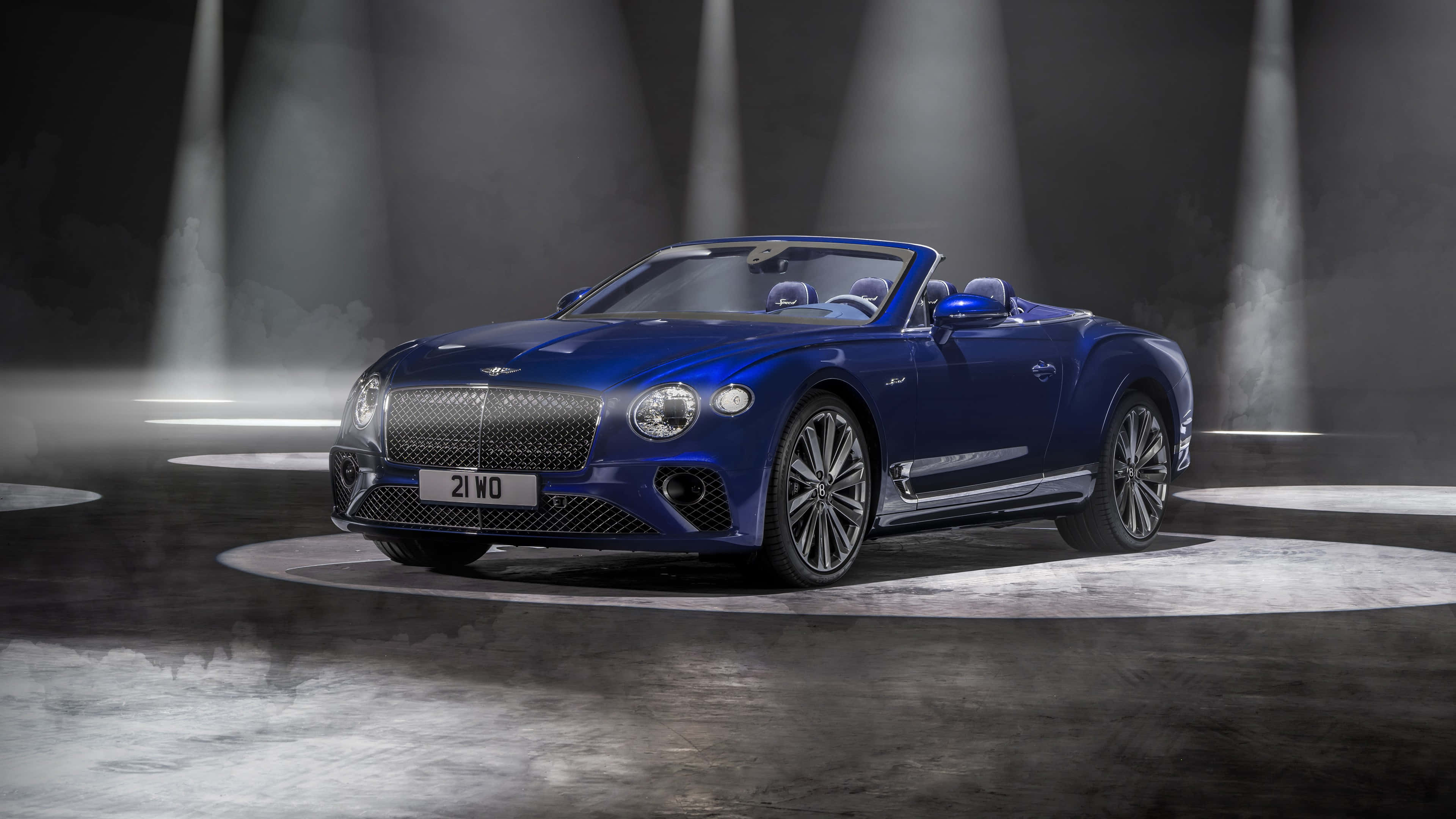 Bentley Continental Gt Convertible 桌布 壁紙 — 一輛時尚的藍色賓利敞篷車在煙霧繚繞的舞台上，在戲劇性的聚光燈下閃閃發光。