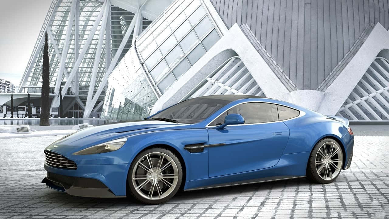 Aston Martin Vanquish 桌布 壁紙 — 一輛時尚的藍色跑車停泊在鵝卵石廣場上，前方是一座未來風格的玻璃和鋼結構建築。
