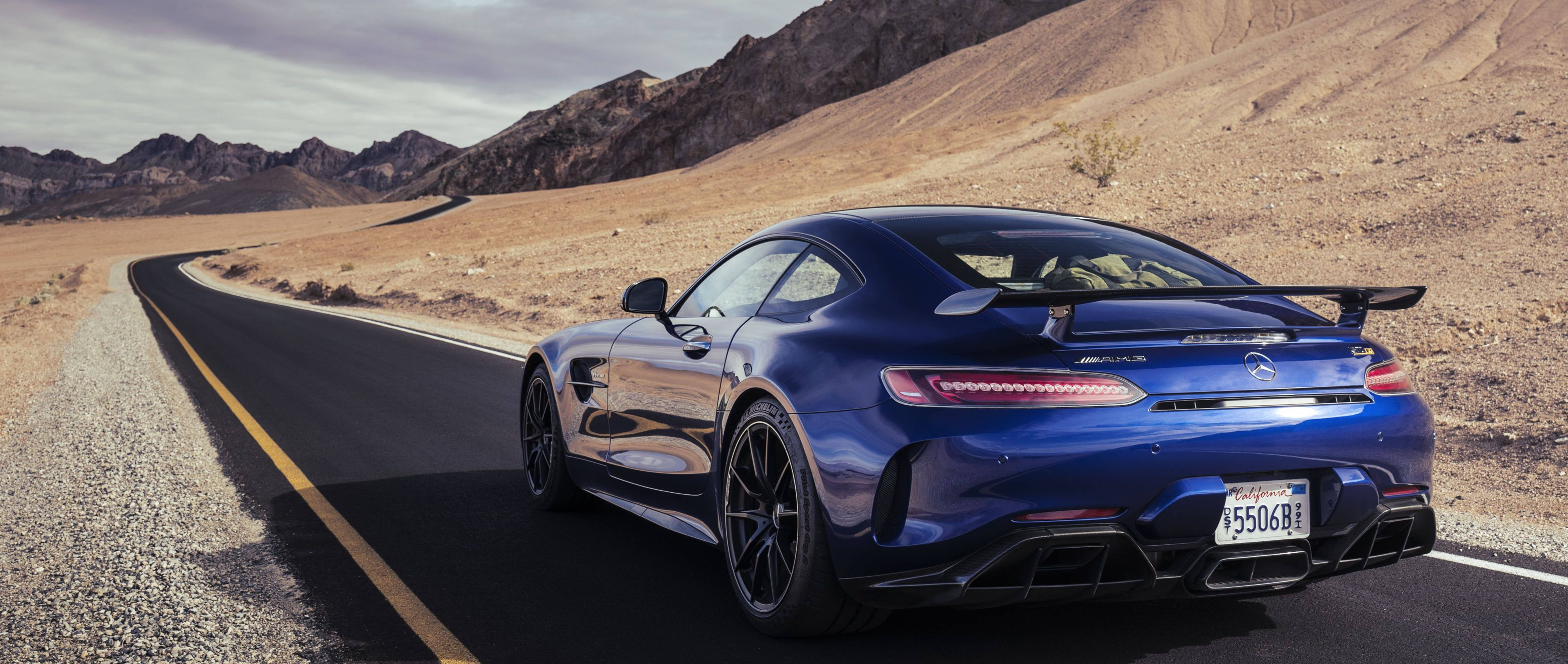 Sports Car 桌布 壁紙 — 一輛時尚的藍色跑車帶有大型後擾流板，在蜿蜒的沙漠公路上行駛，兩旁是崎嶇的山脈，天空多雲。